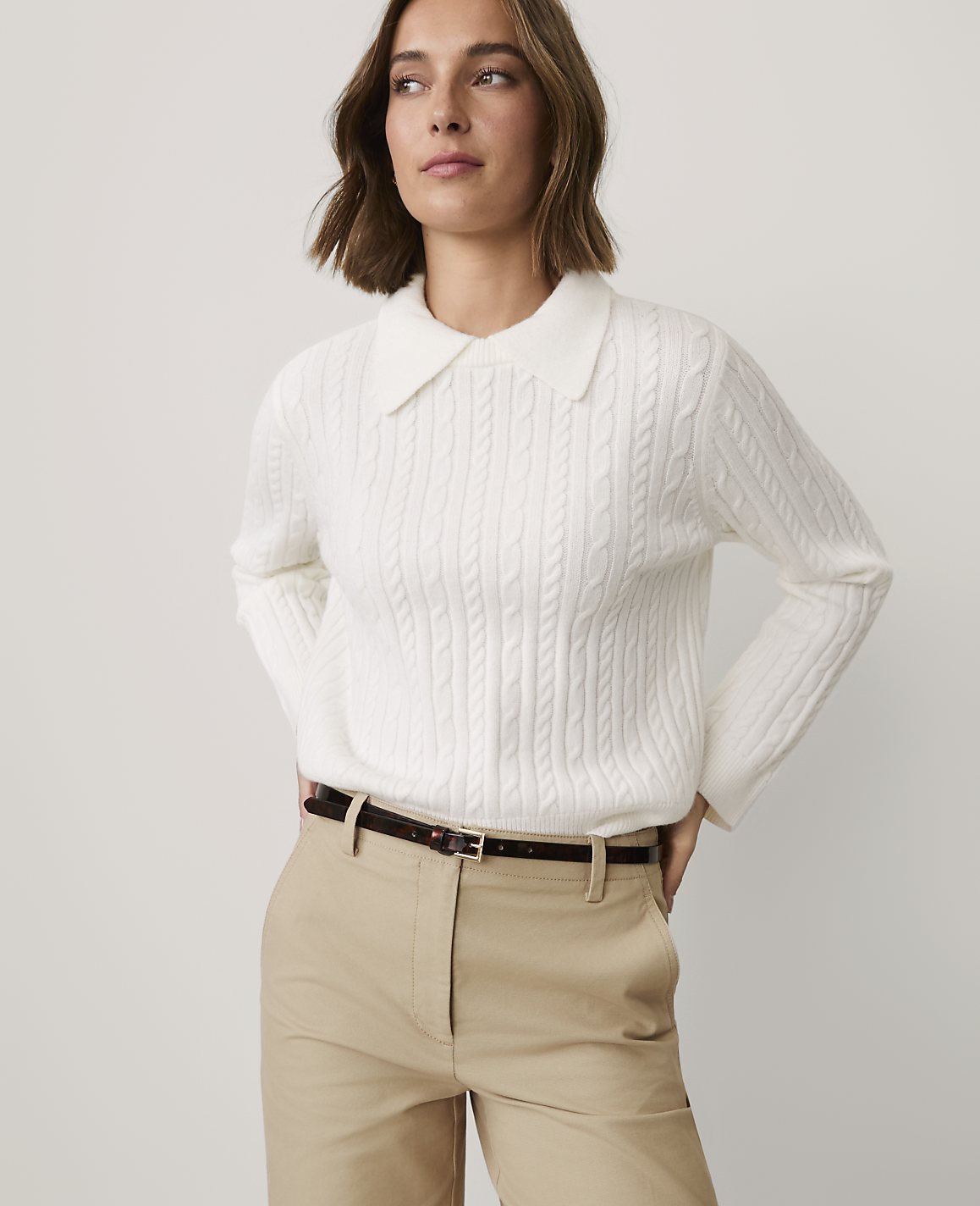 A woman wearing a white cable-knit collared sweater and khaki pants stands against a plain light gray background.