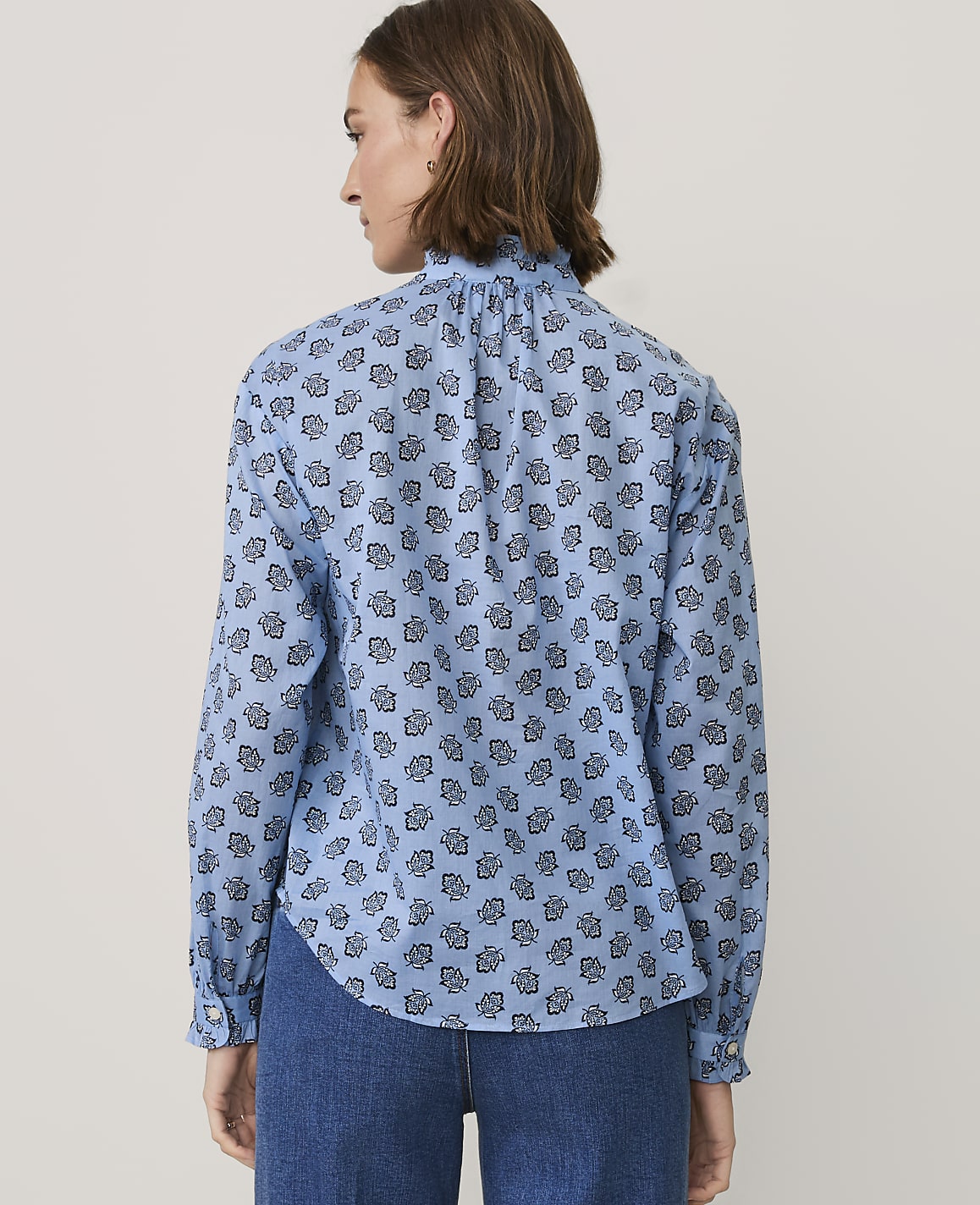 A woman wearing a blue patterned blouse and denim jeans stands against a plain, light-colored background.