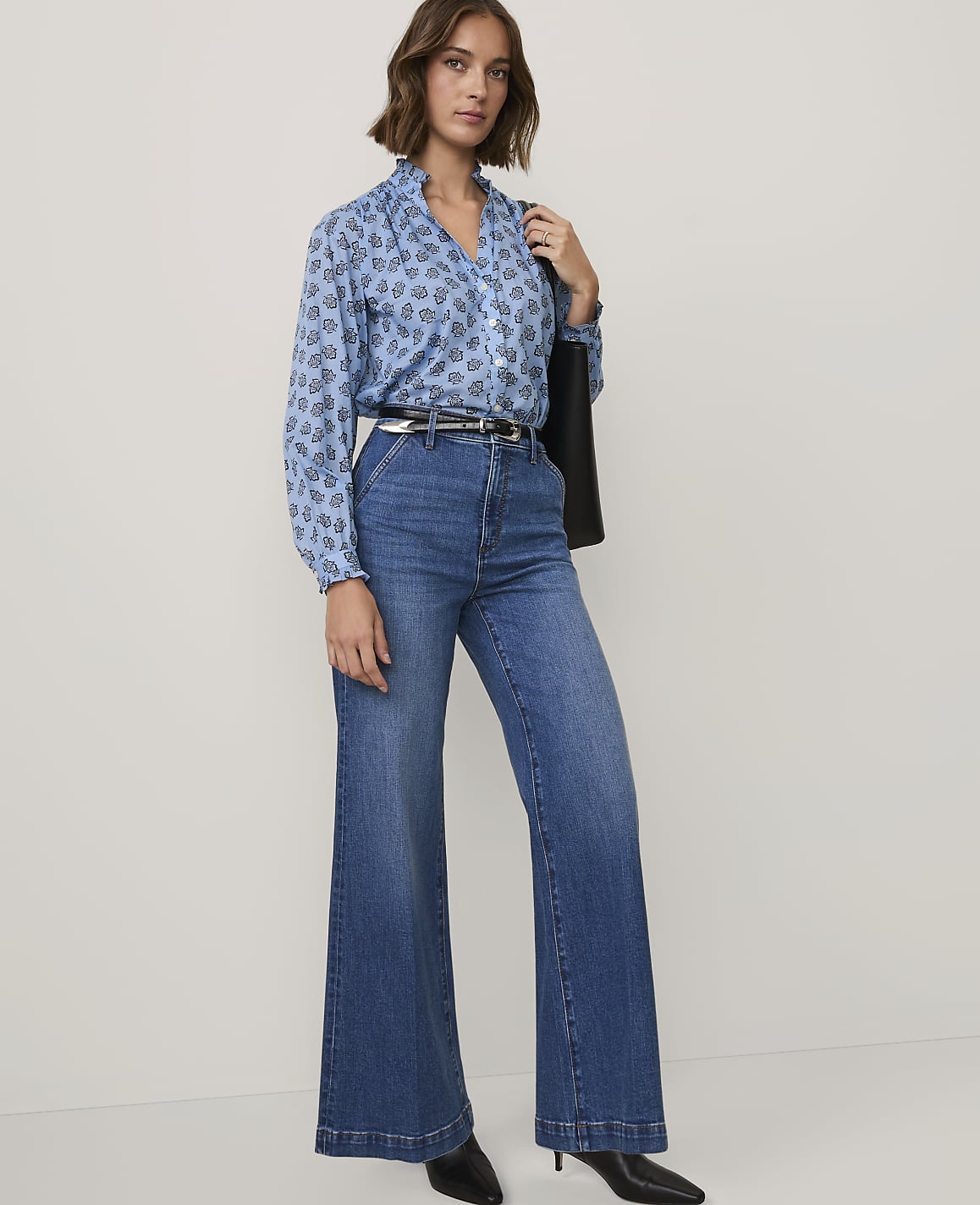 A woman wearing a blue patterned shirt and wide-leg jeans stands against a plain white background.