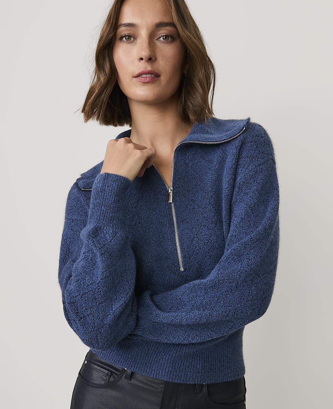 A woman wearing a blue quarter-zip sweater and black leather pants stands against a plain, light-colored background.