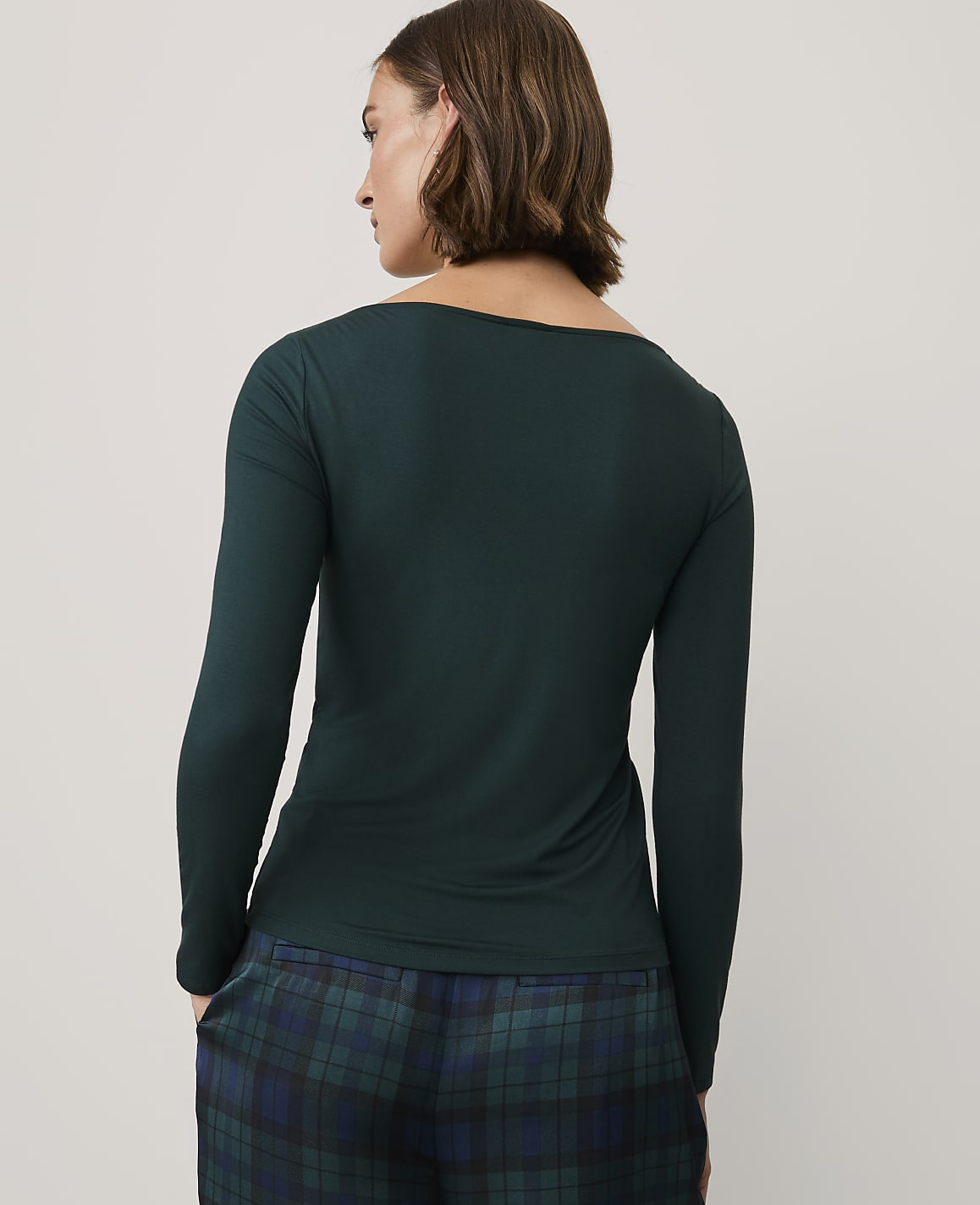 A woman wearing a dark green long-sleeved top and plaid pajama pants stands against a plain light gray background.