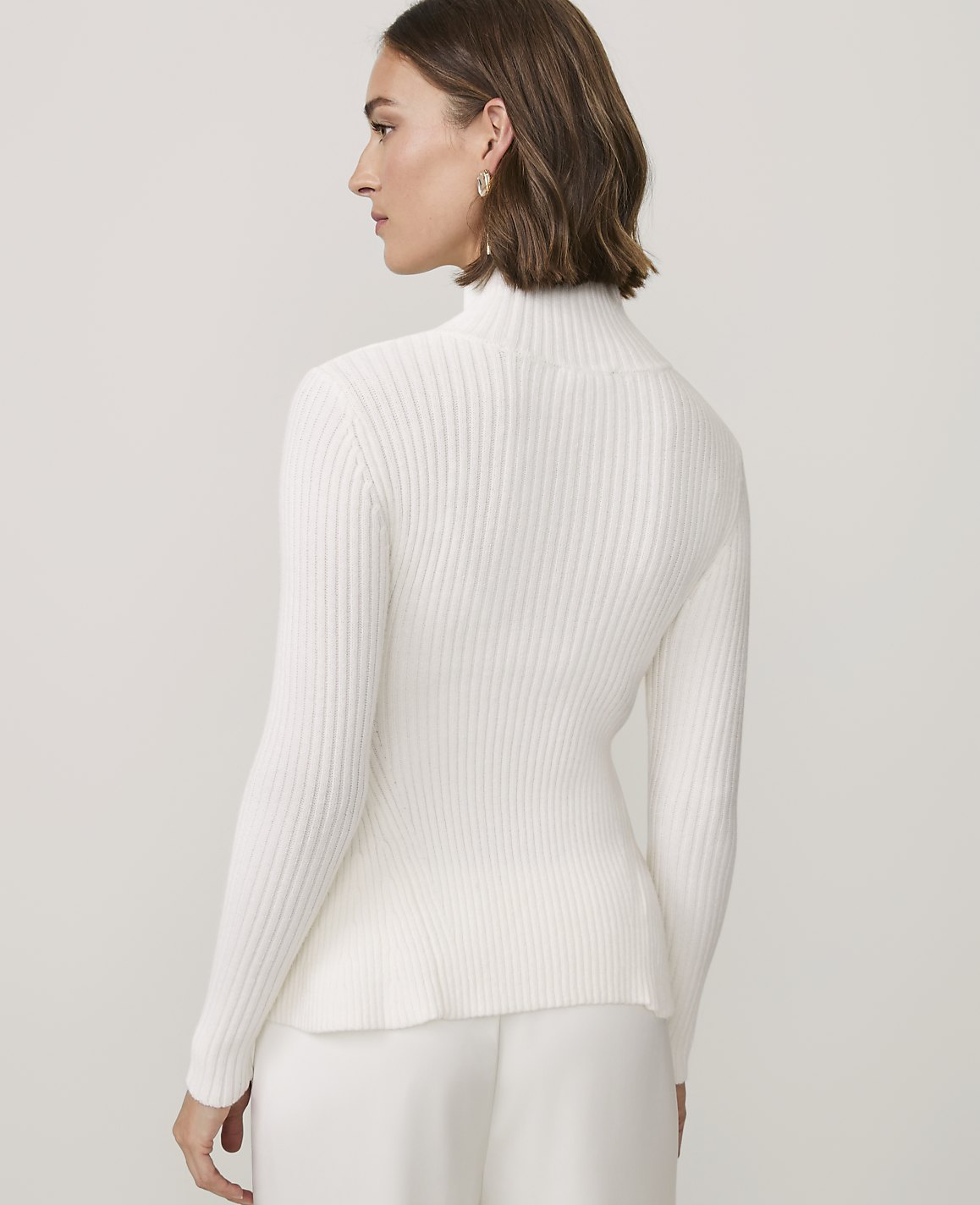 A woman wearing a white ribbed turtleneck sweater and white pants stands against a plain, light-colored background.