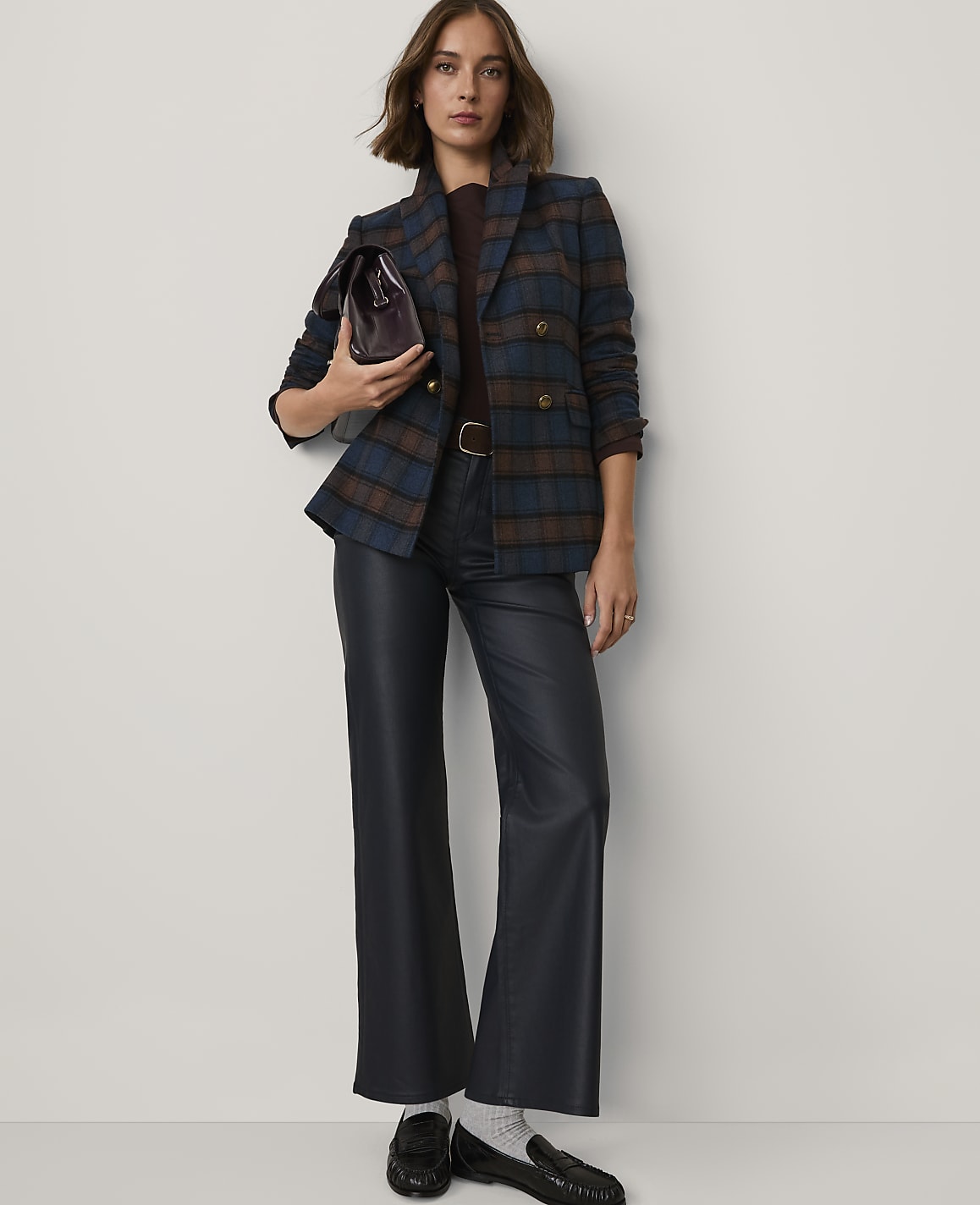 A woman wearing a plaid blazer and navy leather trousers stands against a plain white background.
