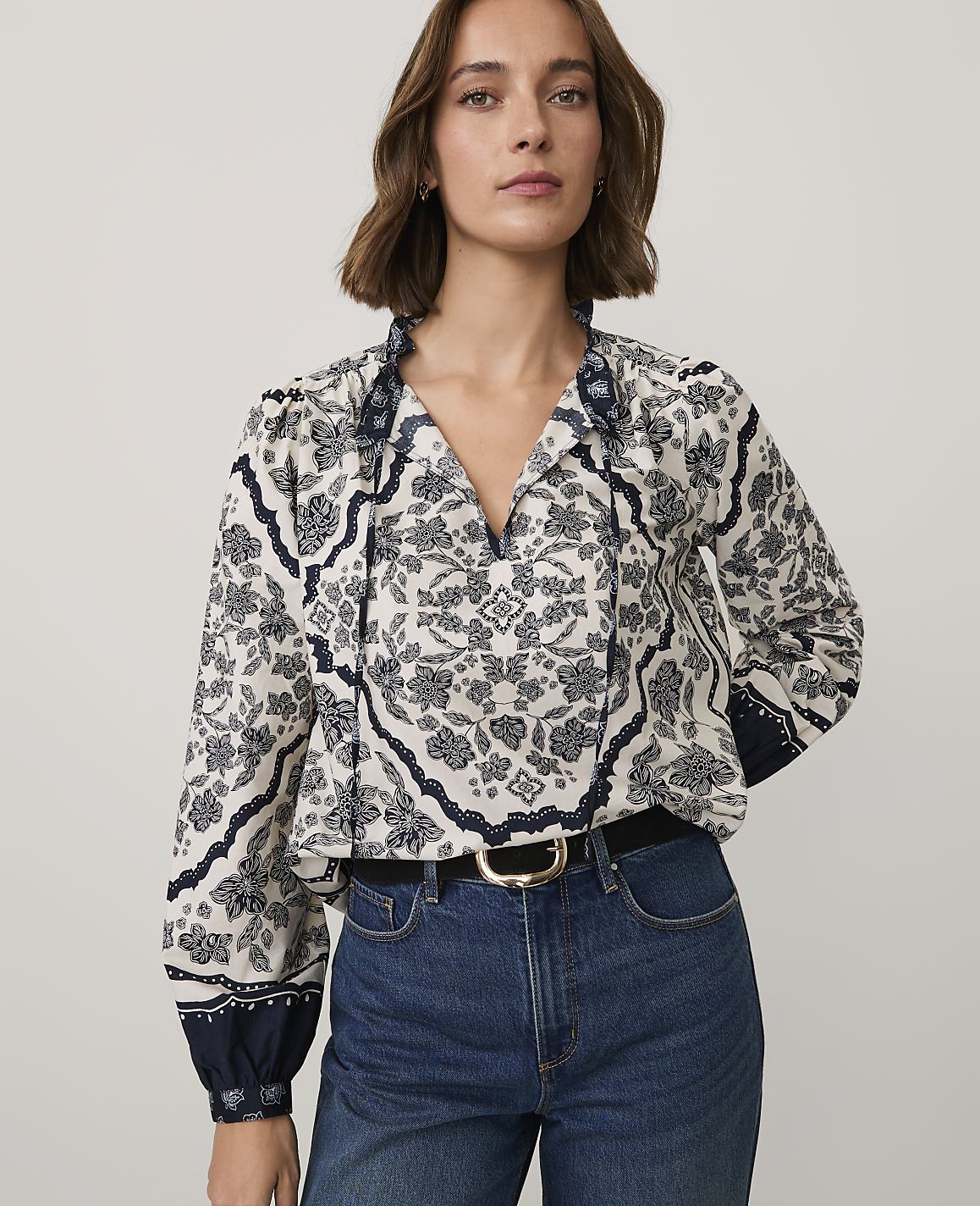 A woman wearing a patterned white and navy blouse and blue jeans stands against a plain light gray background.