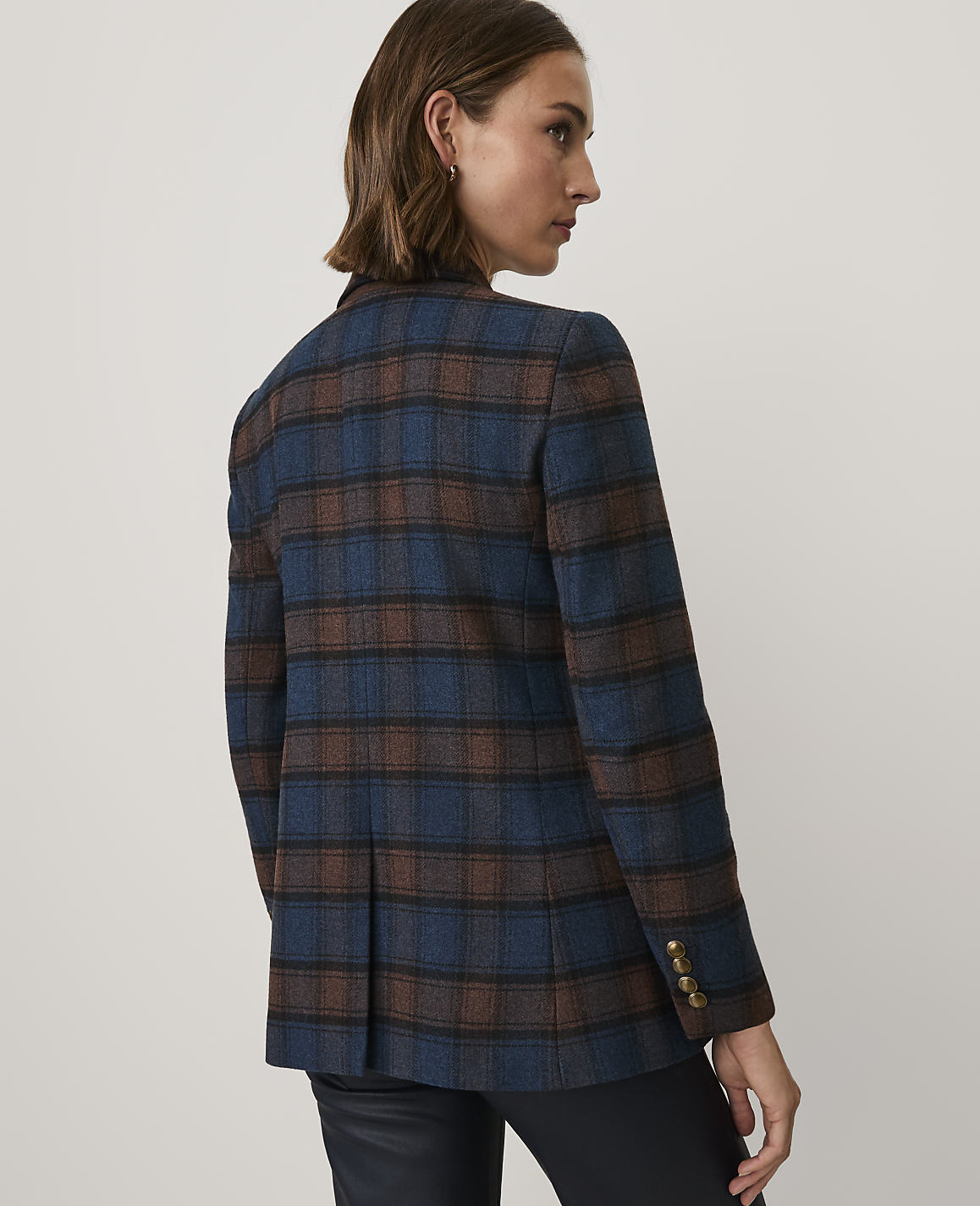 A woman wearing a blue and brown plaid blazer and black pants stands against a plain light gray background.