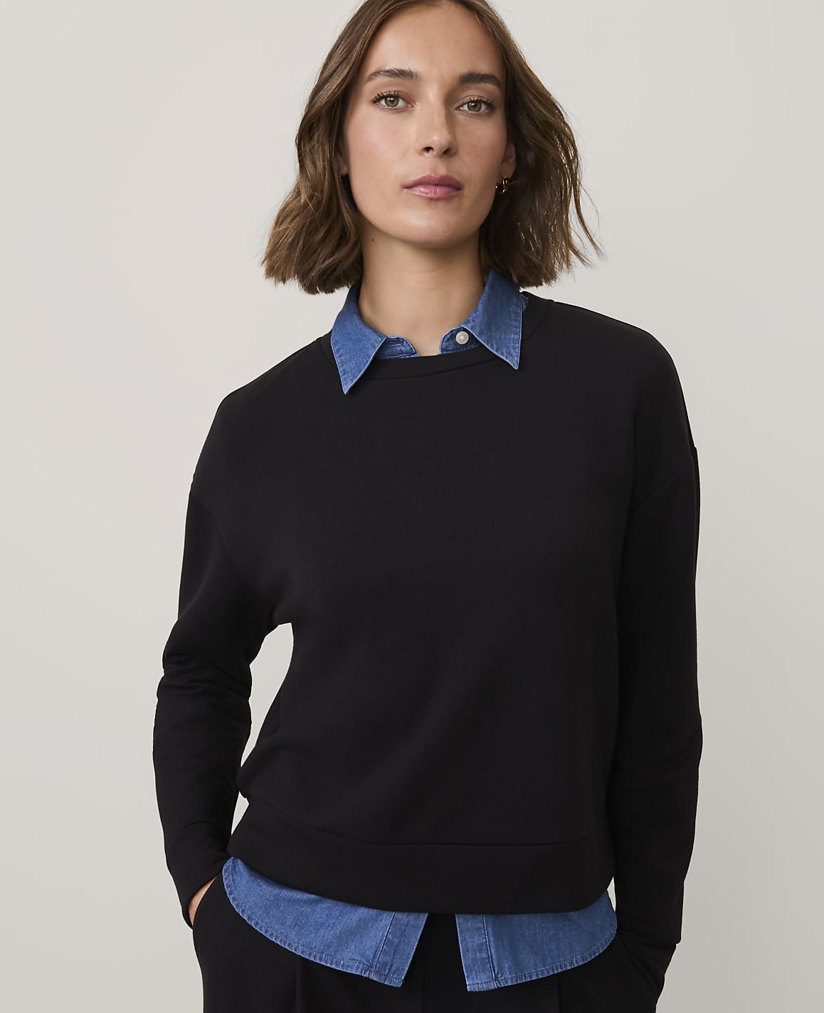 A woman wearing a black sweatshirt over a blue collared shirt stands against a plain, light-colored background.