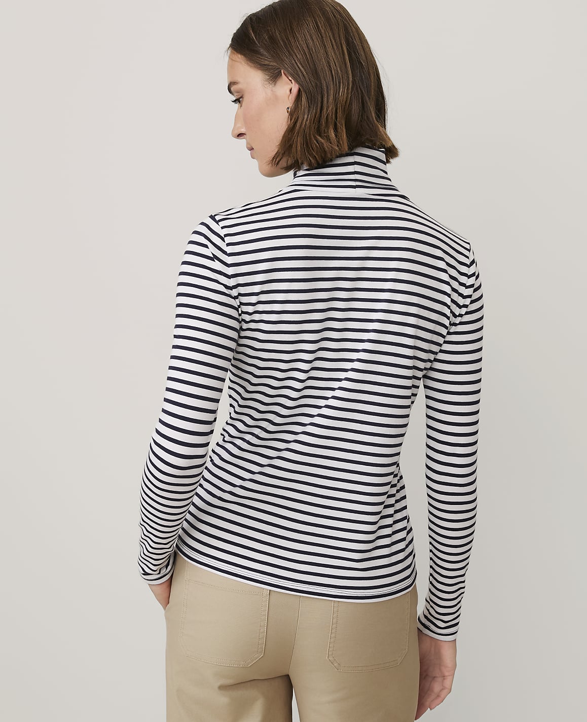 A woman wearing a black and white striped long-sleeved shirt and khaki pants stands against a plain light gray background.