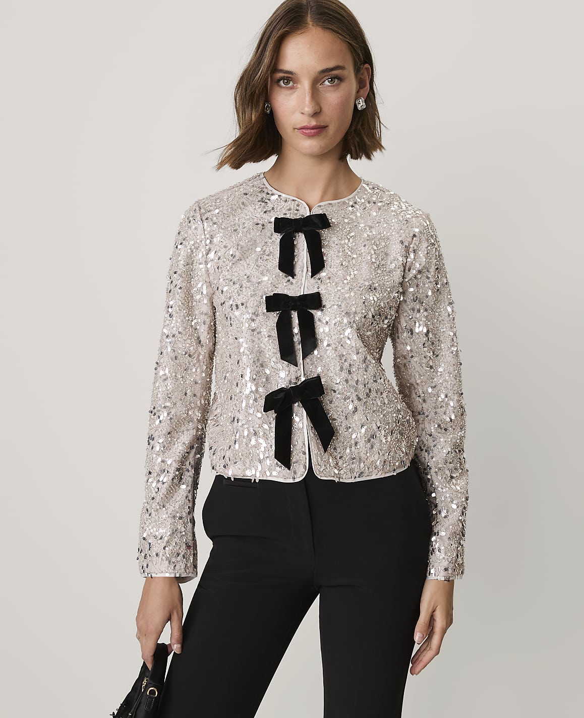 A woman wearing a sequined champagne-colored jacket with black bow accents and black trousers stands against a plain light gray background.
