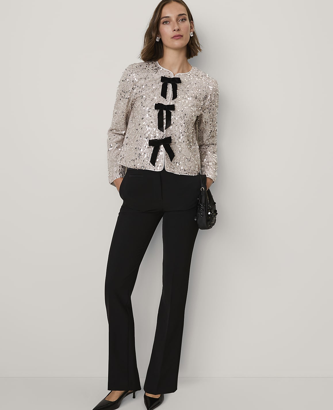 A woman wearing a sequined jacket with black bow accents and black trousers stands against a plain, light-colored background.