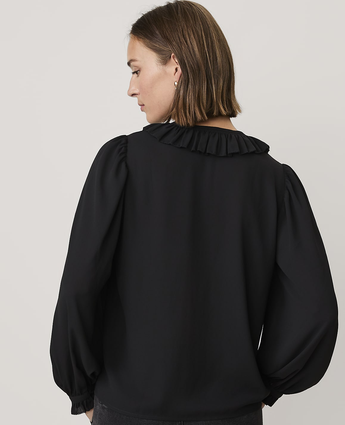 A woman wearing a black ruffled blouse stands with her back to the camera against a plain light gray background.