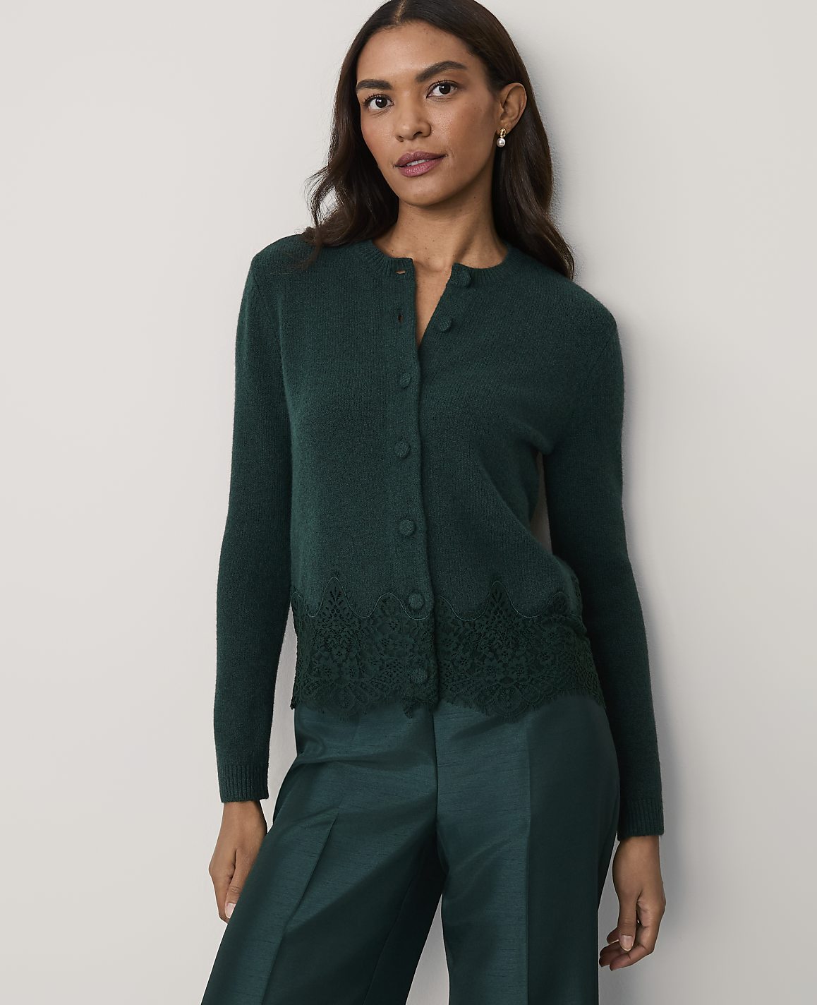 A woman wearing a dark green cardigan with lace detailing and matching trousers stands against a plain, light-colored wall.