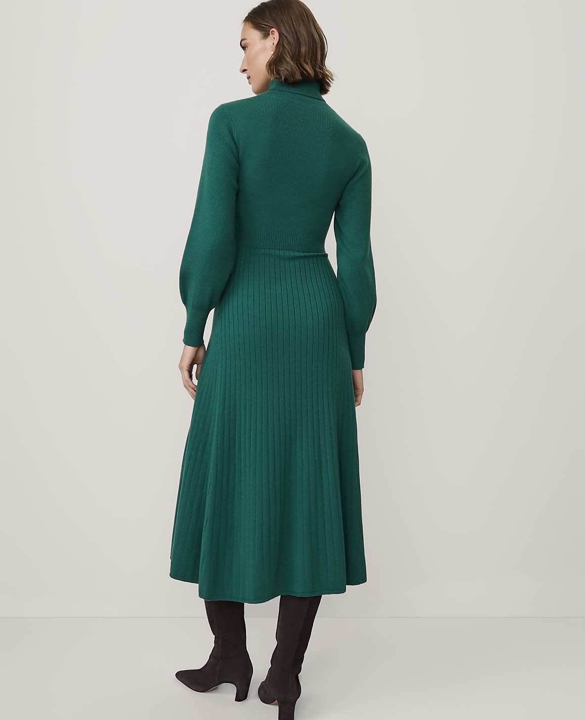 A woman wearing a long-sleeved green ribbed knit dress and dark brown boots stands against a plain white background.