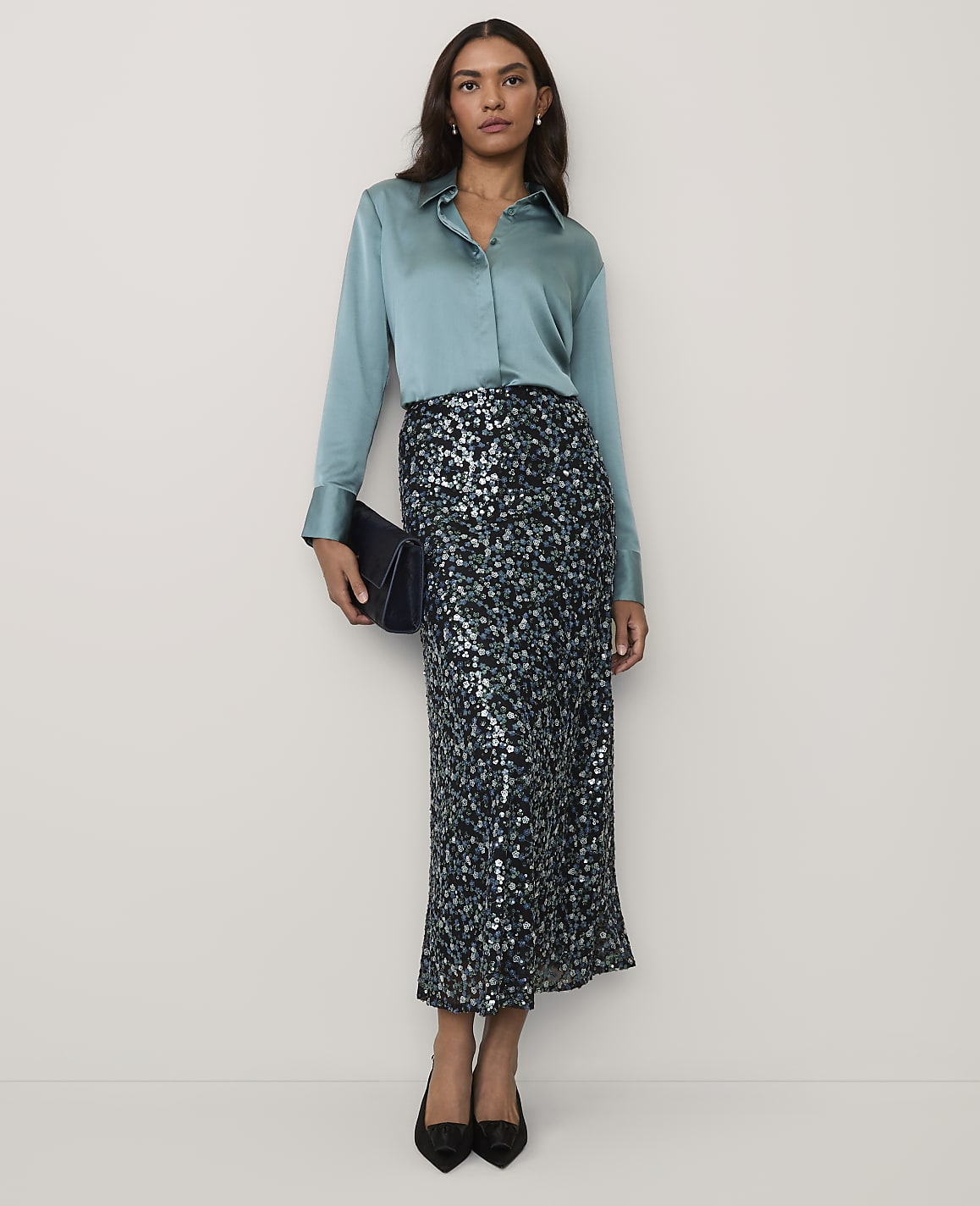 A woman wearing a light blue satin shirt and a sequined maxi skirt stands against a plain, neutral background.
