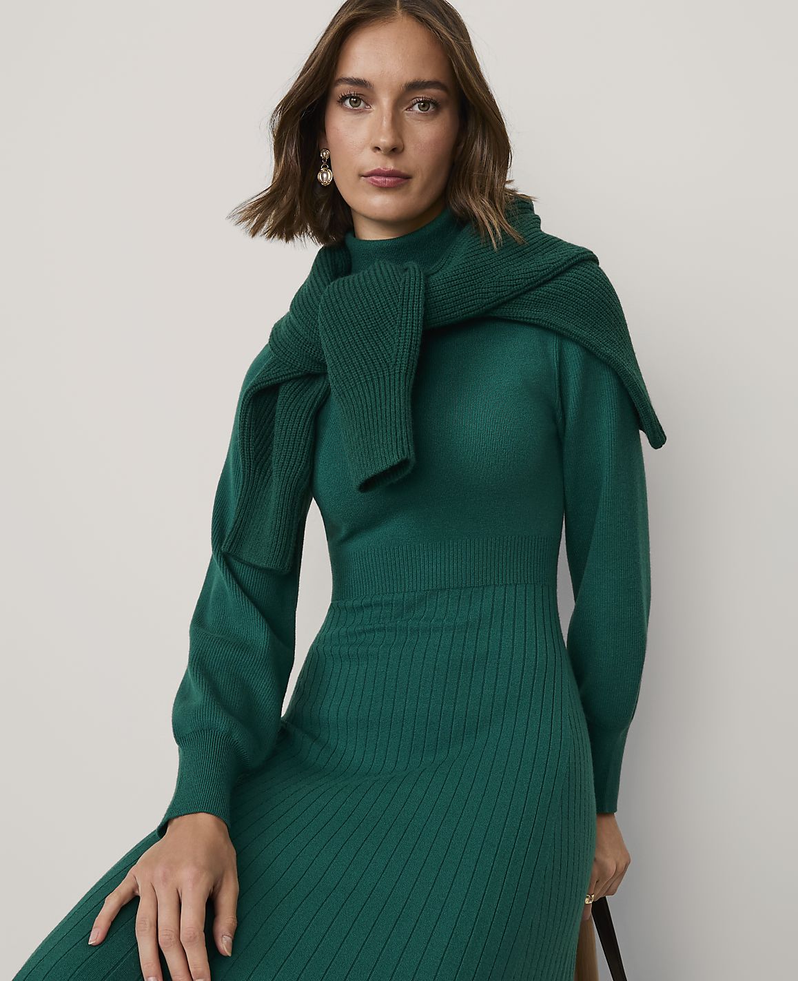 A woman wearing a green ribbed knit dress and a matching sweater draped over her shoulders poses against a plain light gray background.