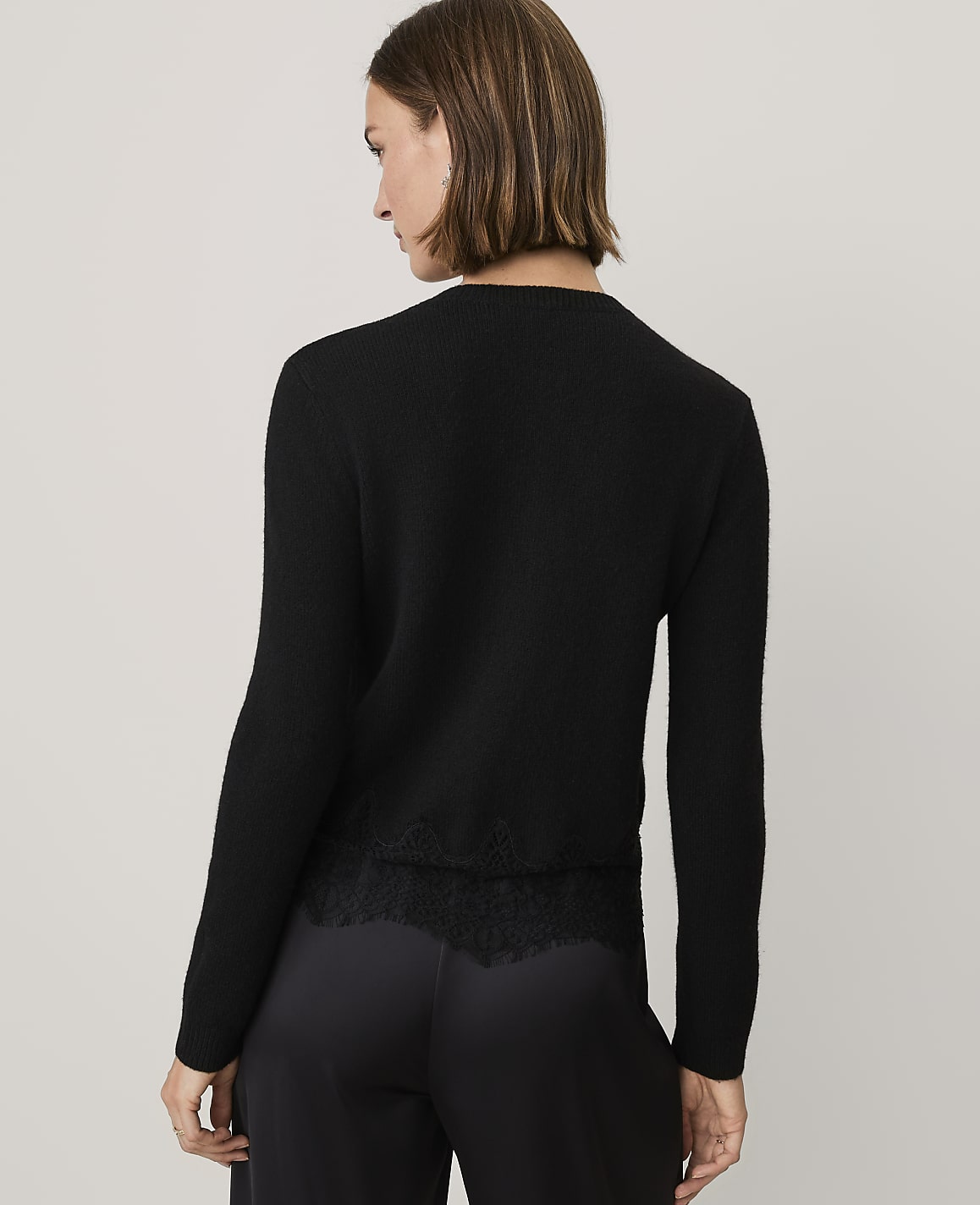A woman wearing a black sweater with lace detailing and black trousers stands against a plain light gray background.