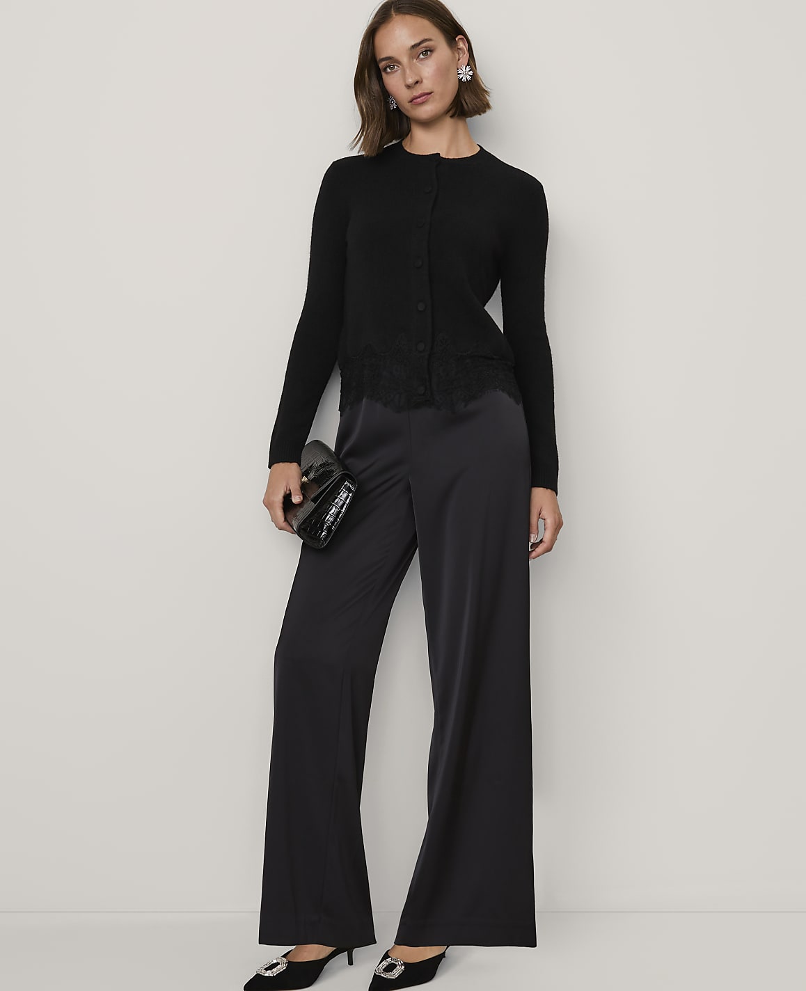 A woman wearing a black cardigan and wide-leg satin trousers stands against a plain white background.