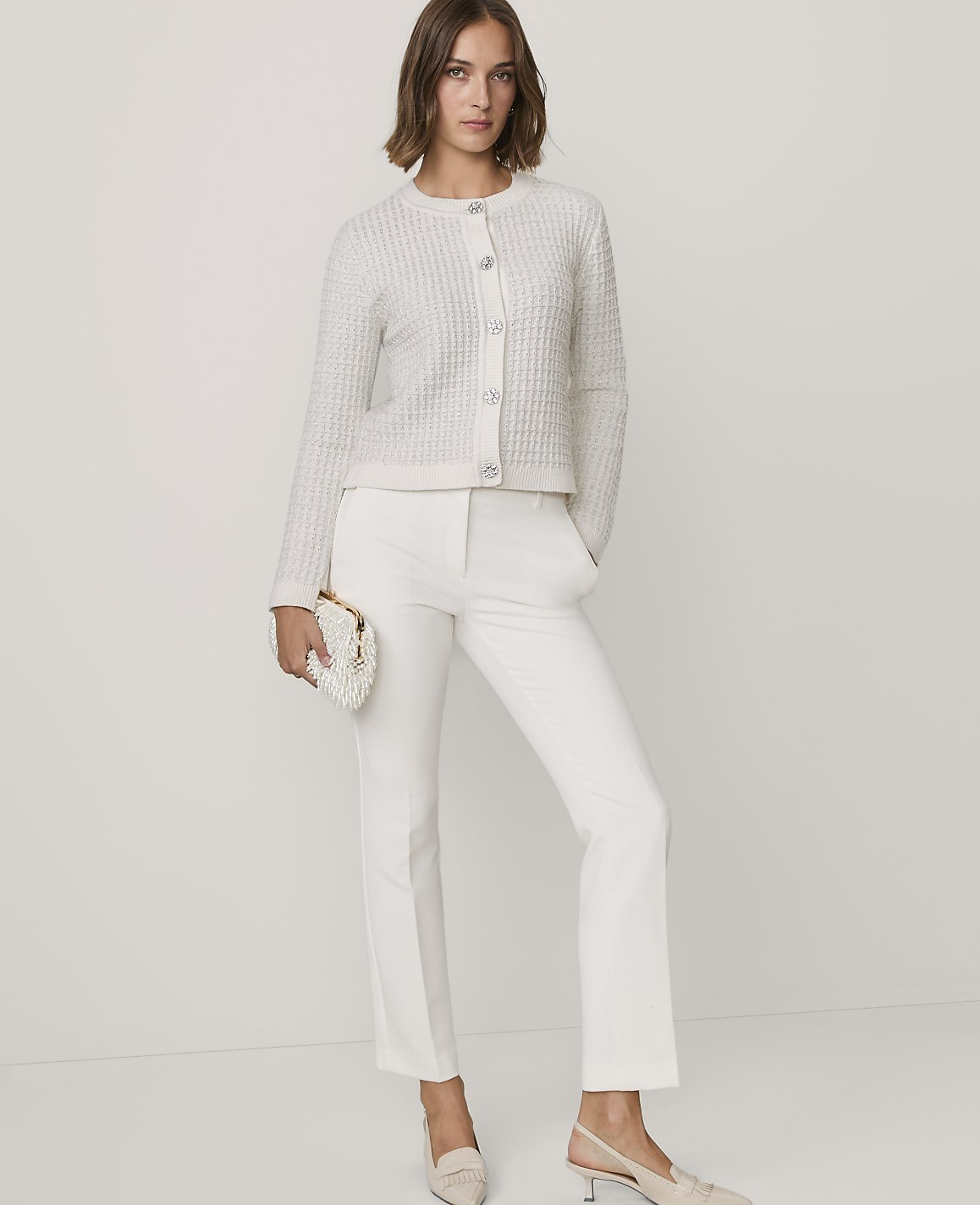 A woman wearing a white textured cardigan, white trousers, and beige heels stands against a plain white background.