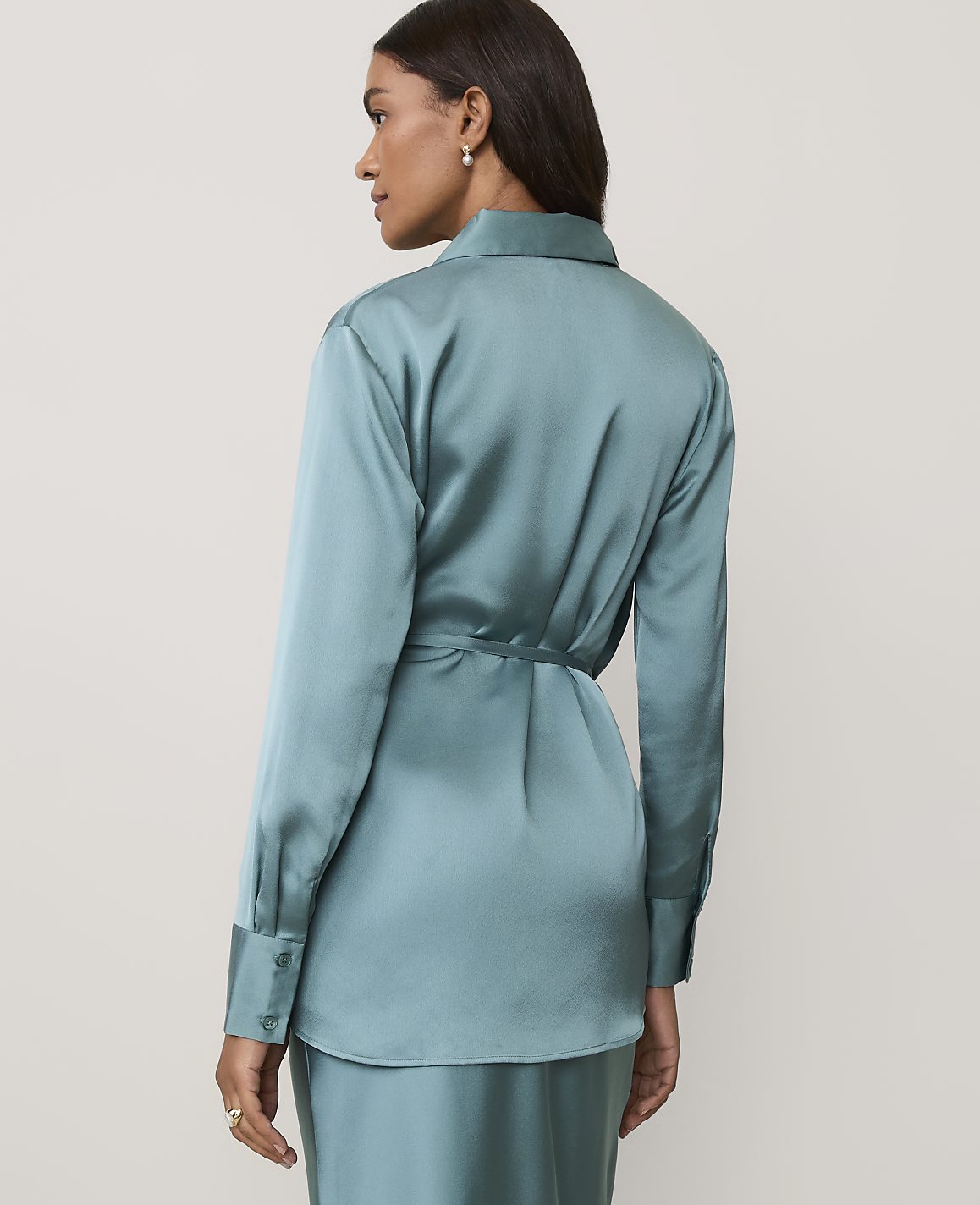 A woman wearing a teal satin shirt and matching trousers stands against a plain, light-colored background.