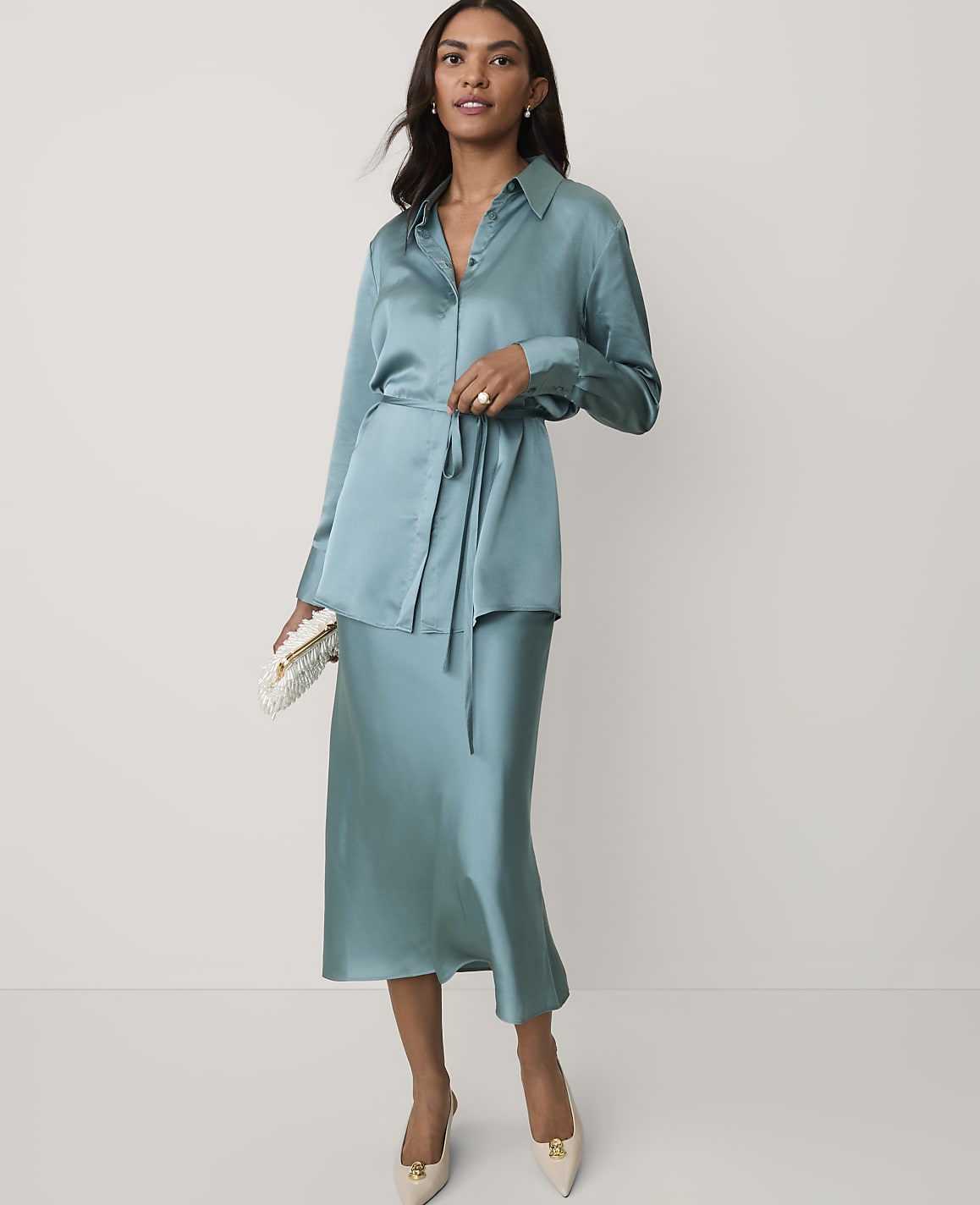 A woman wearing a teal satin shirt and matching midi skirt stands against a plain white background.