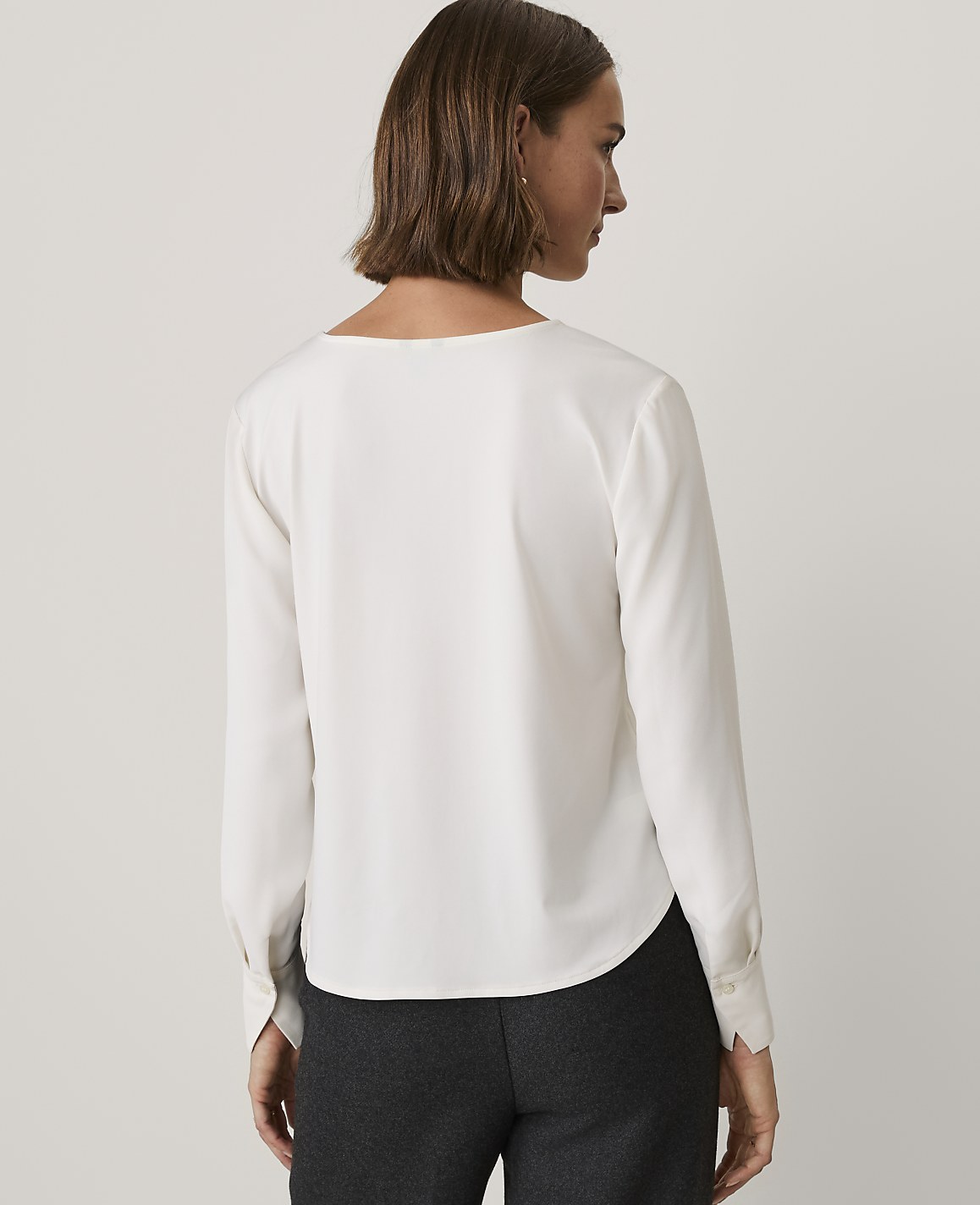 A woman wearing a white long-sleeved blouse and dark trousers stands with her back to the camera against a plain light gray background.