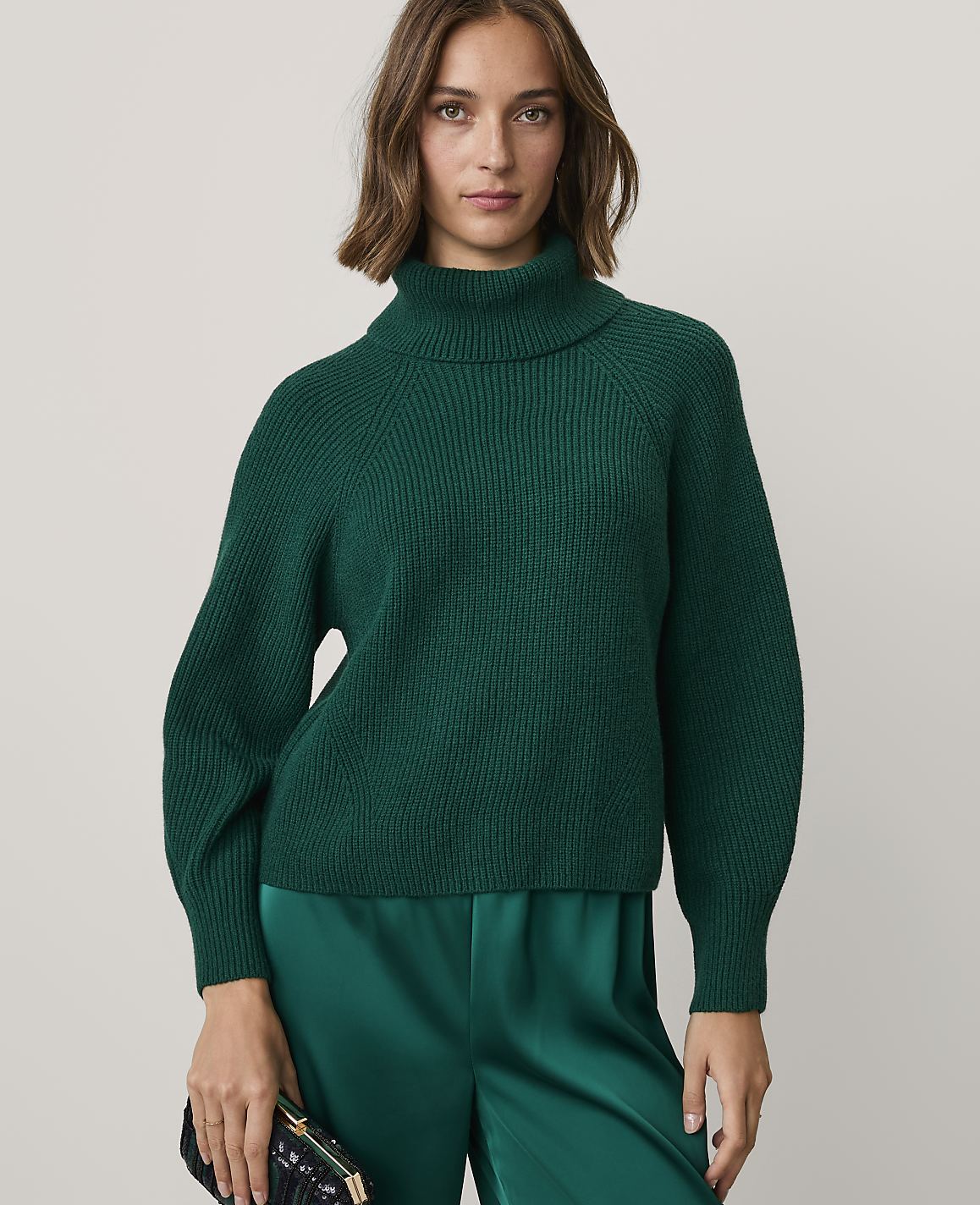 A woman wearing a dark green ribbed turtleneck sweater and matching satin trousers stands against a plain, light-colored background.