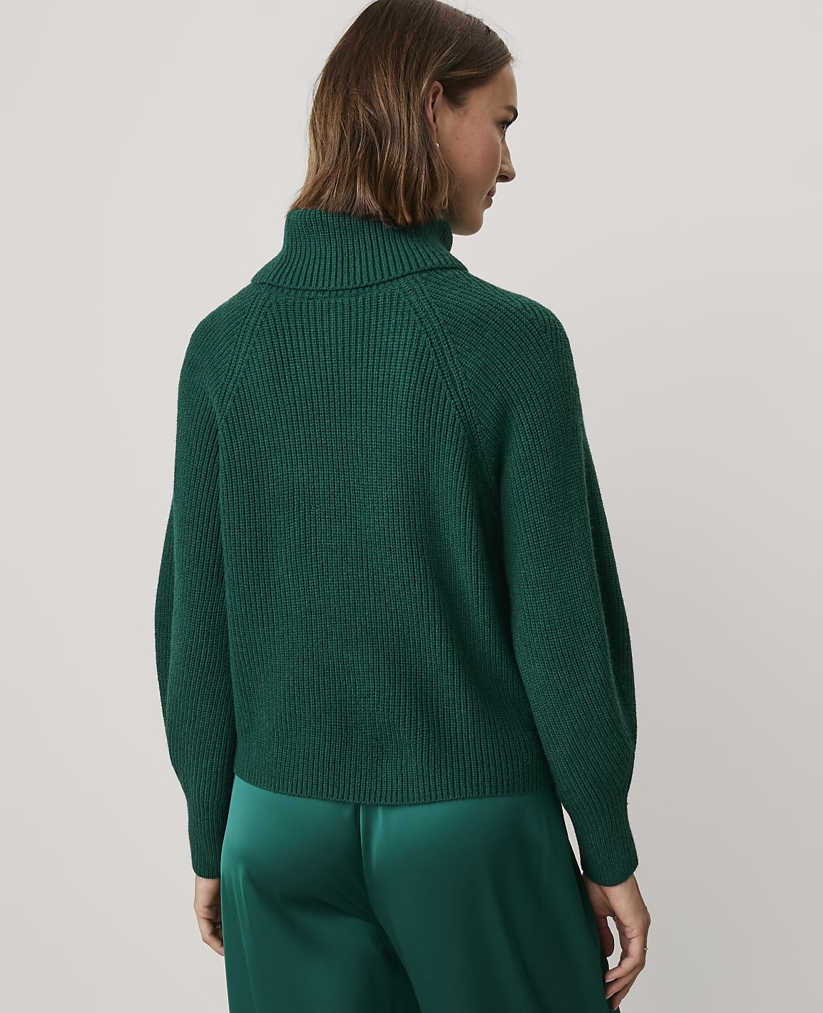 A woman wearing a dark green ribbed turtleneck sweater and matching satin trousers stands against a plain, light-colored background.