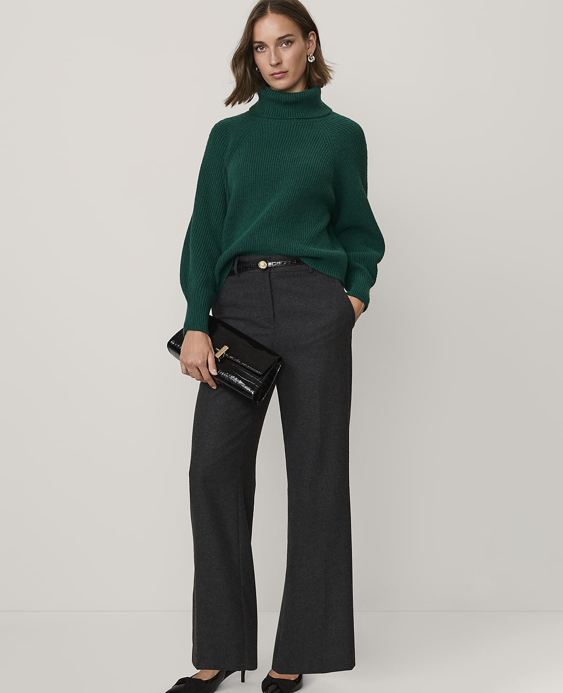 A woman wearing a green turtleneck sweater and grey wide-leg trousers stands against a plain white background.