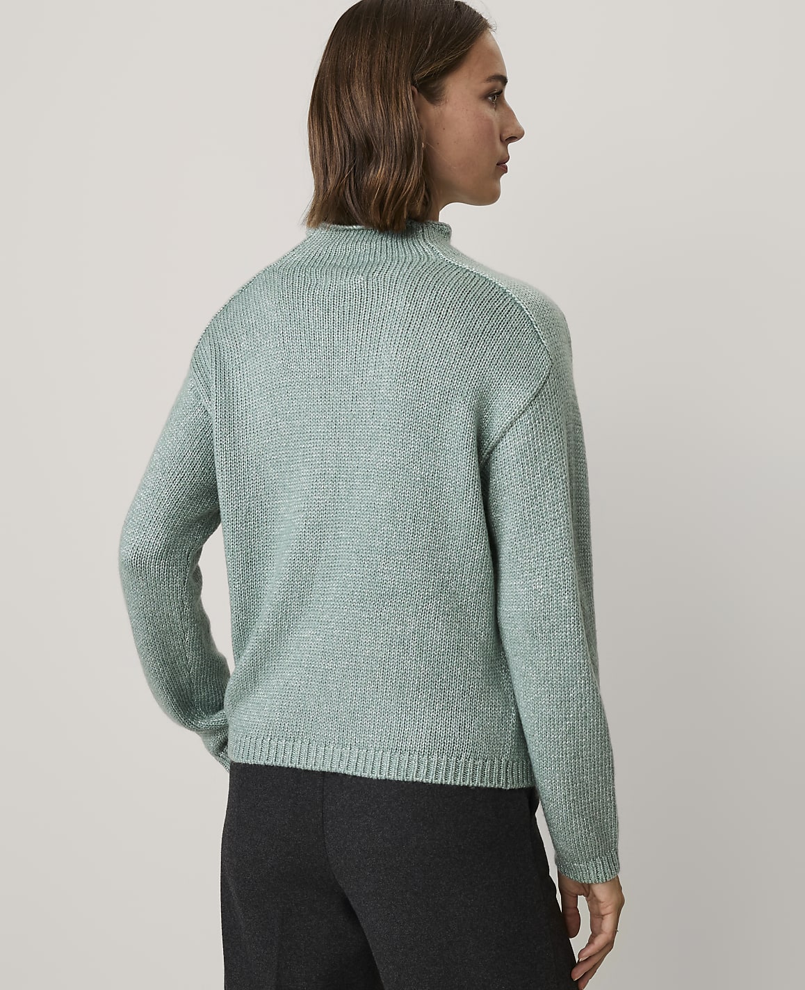 A woman wearing a light green knit sweater and dark trousers stands against a plain light gray background.
