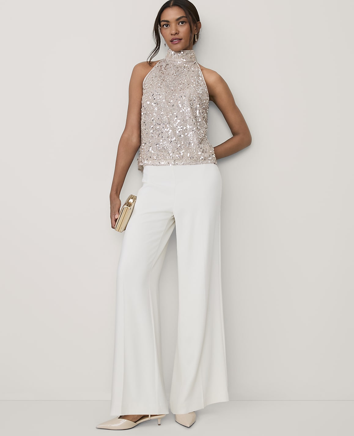 A woman wearing a sequined gold top and wide-leg white trousers stands against a plain, light-colored background.