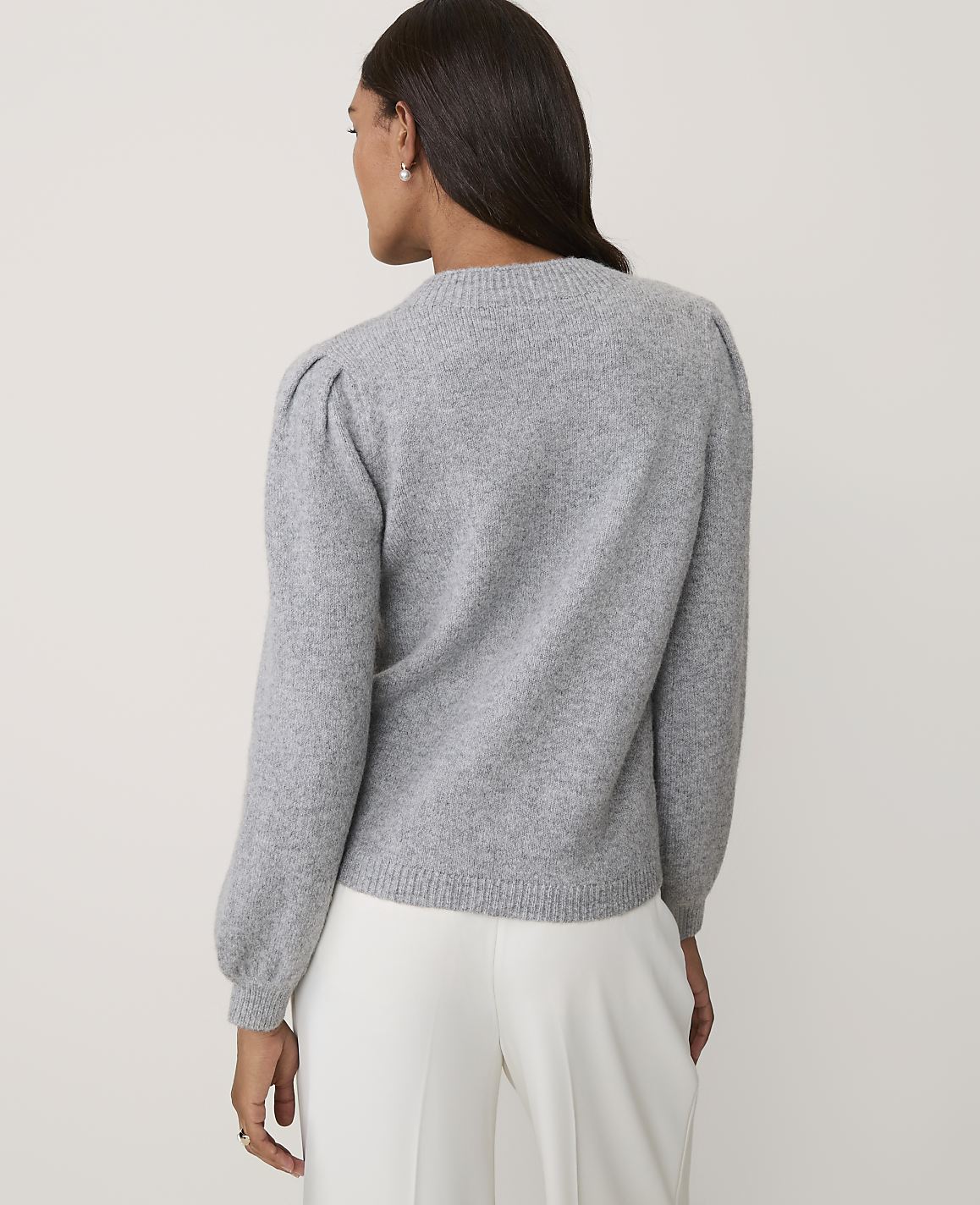 A woman wearing a grey puff-sleeved sweater and white trousers stands against a plain, light-colored background.