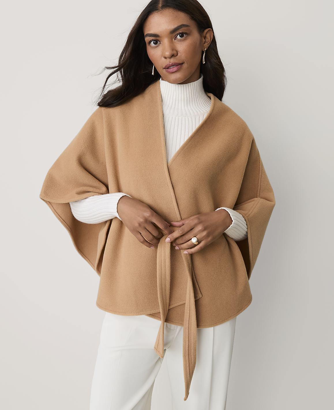 A woman wearing a tan wrap over a white turtleneck and trousers stands against a plain light gray background.