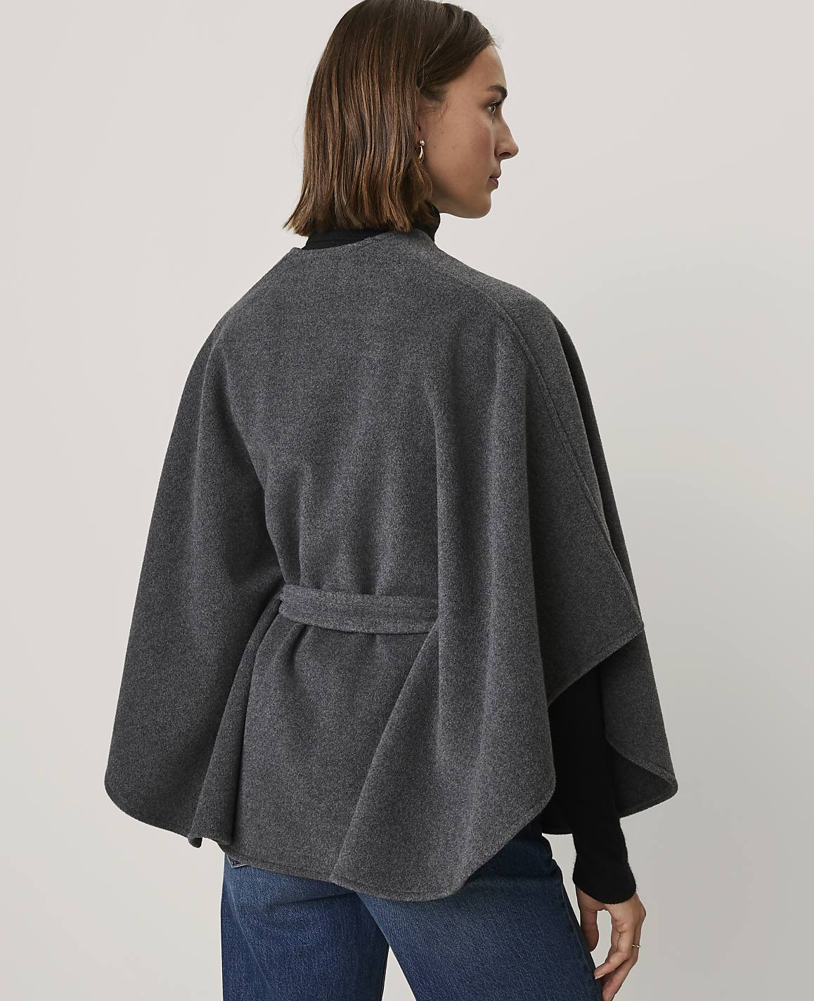 A woman wearing a grey cape and blue jeans stands against a plain white background.