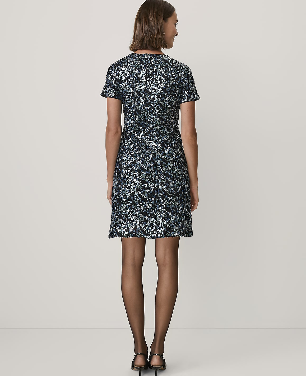 A woman stands with her back to the camera wearing a short, sequined blue and black dress and black heels against a plain white background.