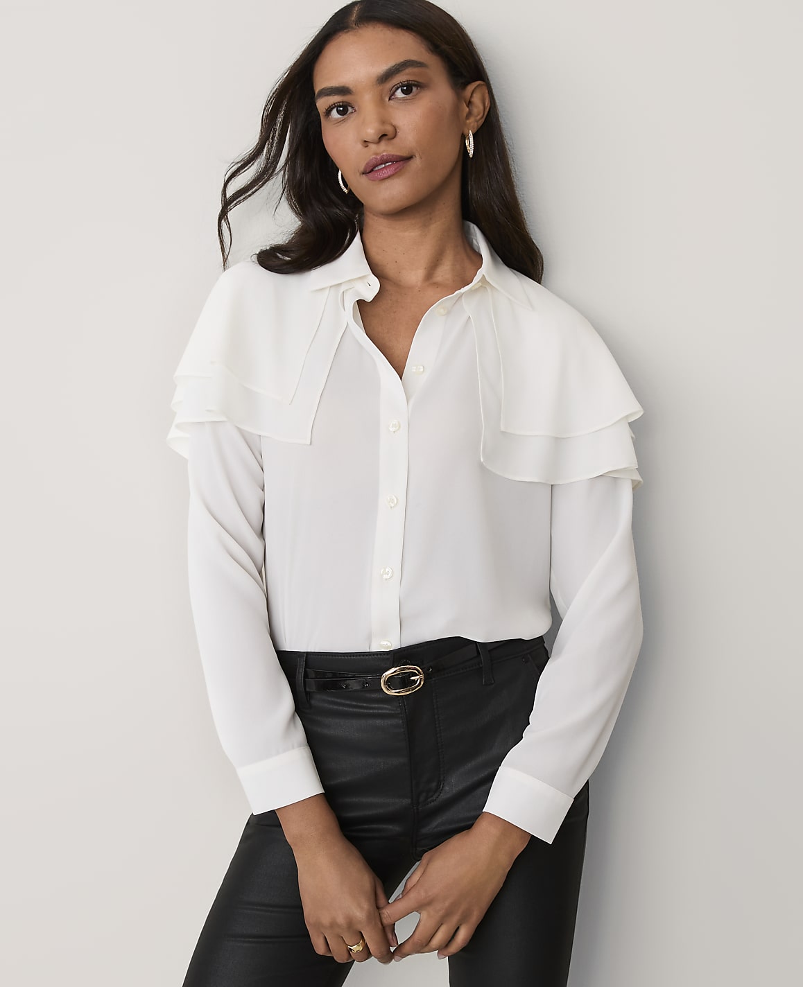 A woman wearing a white ruffled blouse and black leather pants stands against a plain light-grey background.