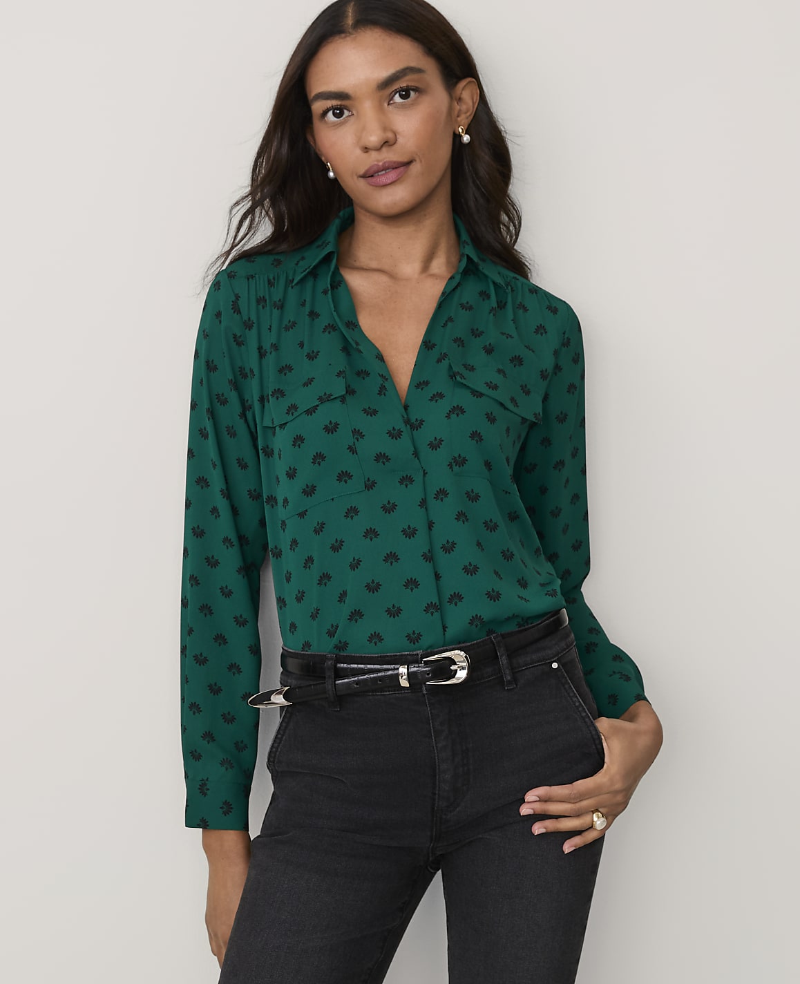 A woman wearing a green patterned button-down shirt and black jeans stands against a plain white background.