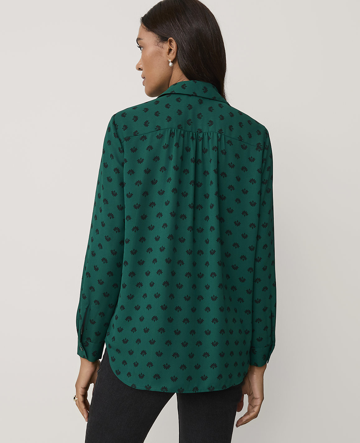 A woman wearing a green patterned shirt and black trousers stands against a plain, light-colored background.