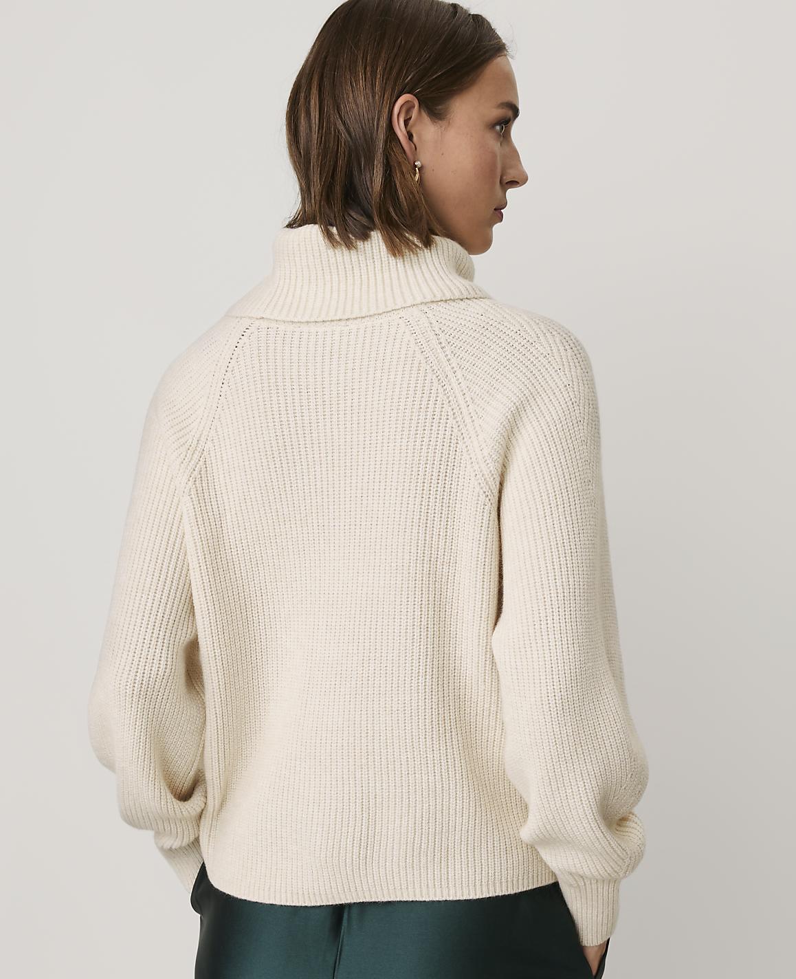 A woman wearing a cream-colored ribbed turtleneck sweater and dark green trousers stands against a plain light gray background.