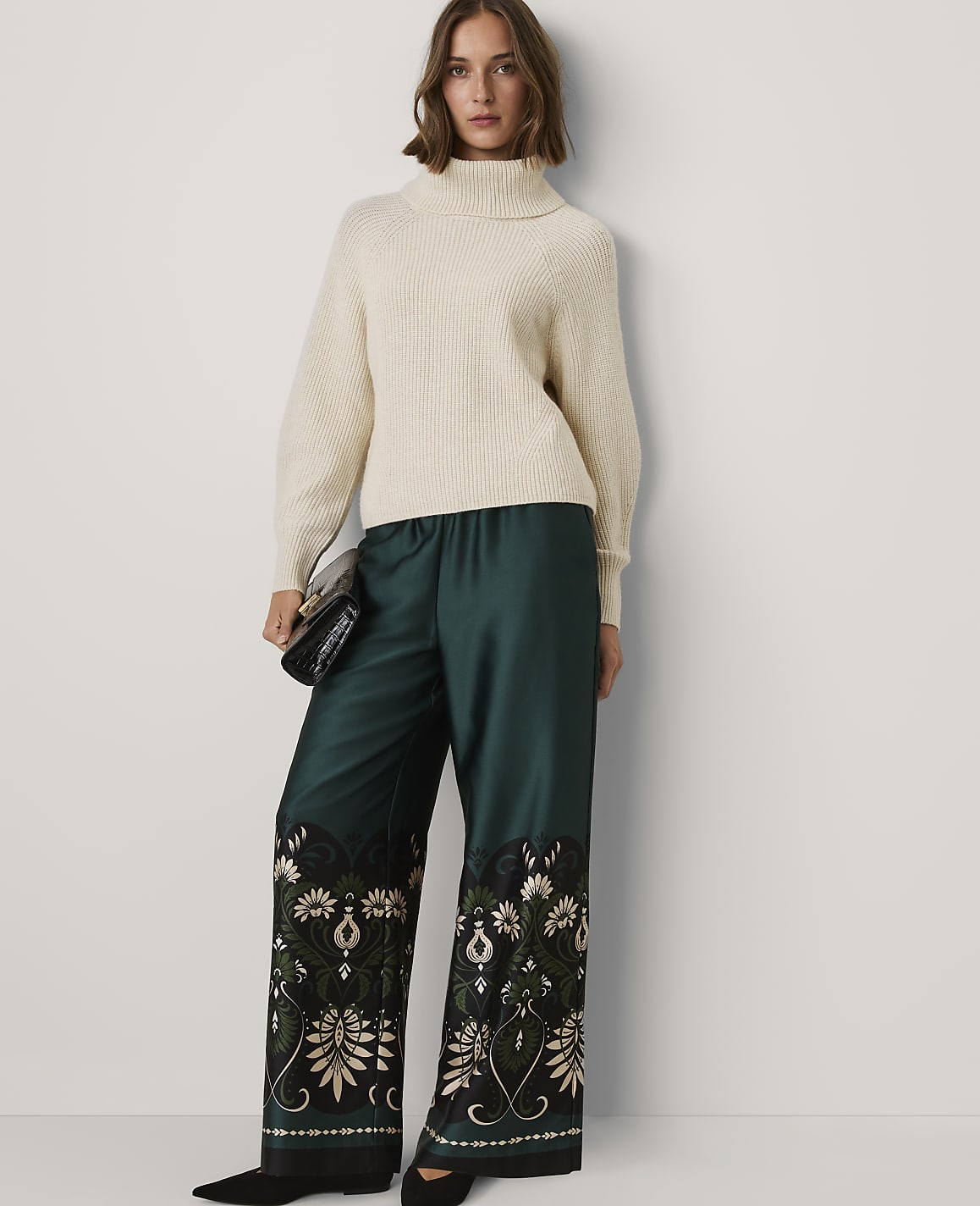 A woman wearing a cream turtleneck sweater and wide-leg teal trousers with floral embroidery stands against a plain white wall.