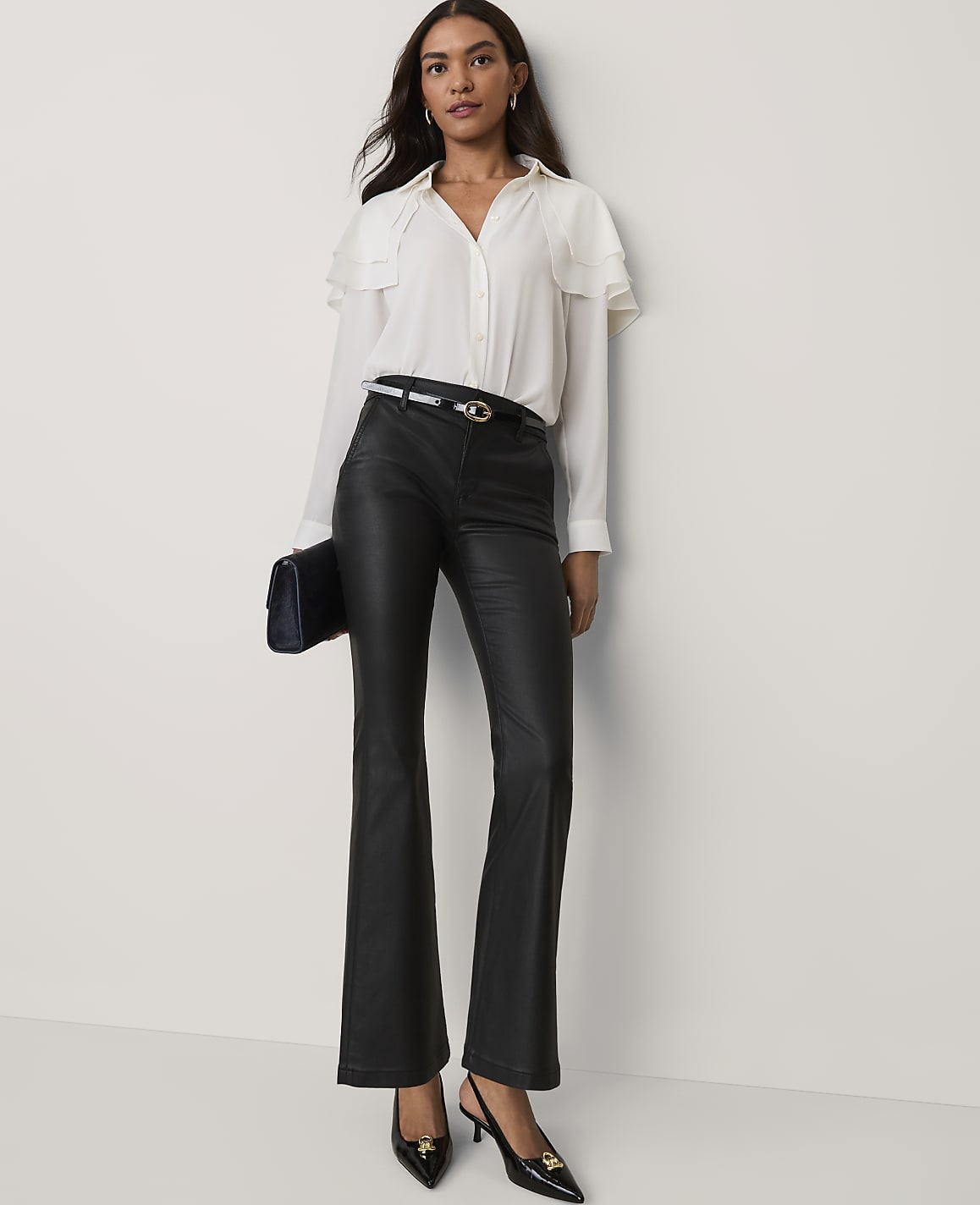 A woman wearing a white ruffled blouse and black faux-leather trousers stands against a plain white wall.
