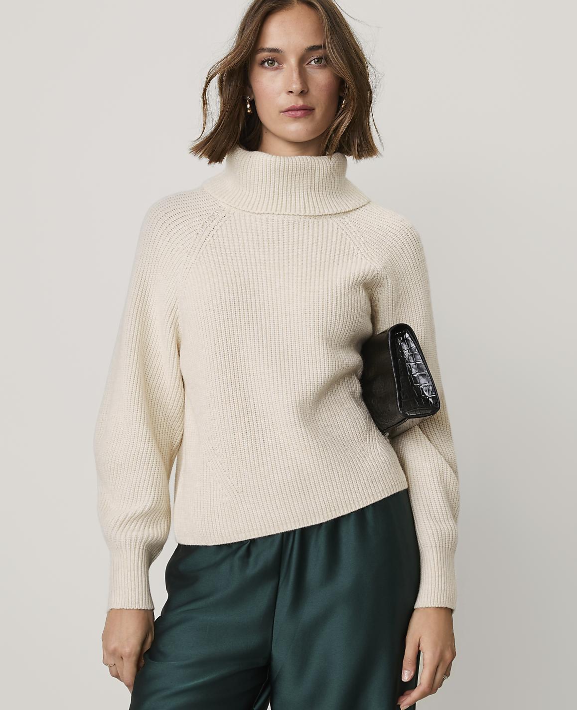 A woman wearing a cream-colored turtleneck sweater and dark green satin pants holds a black clutch against a plain light gray background.