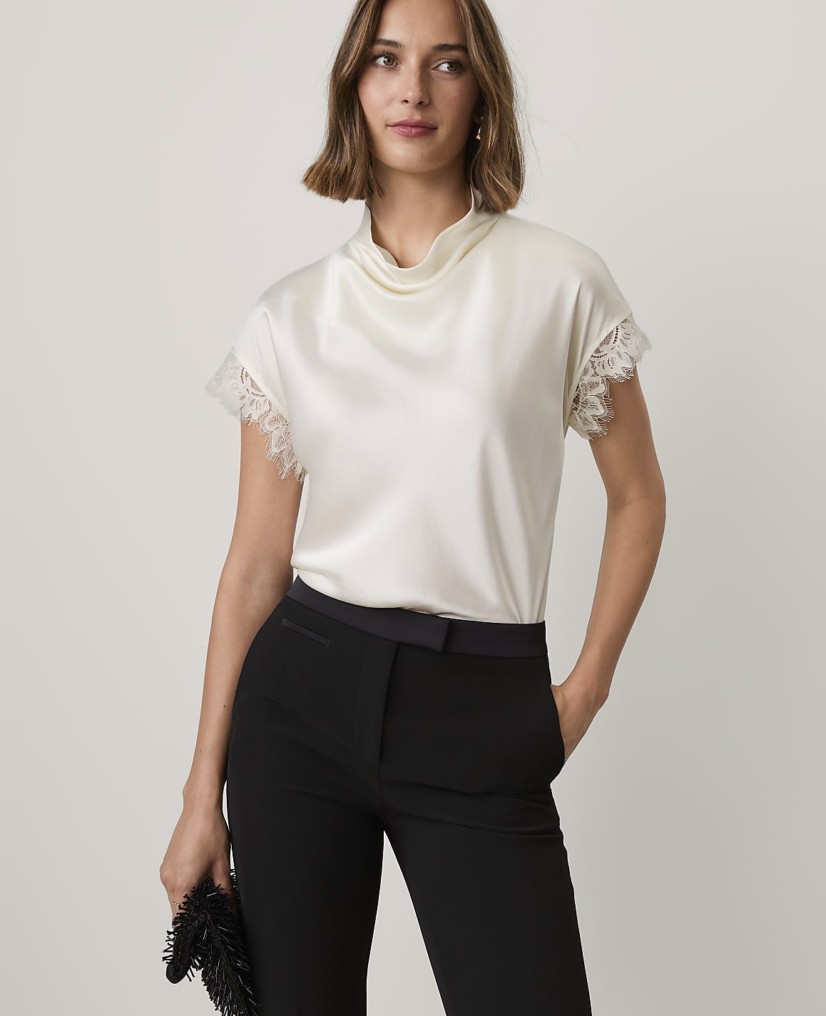 A woman wearing a white satin top and black trousers stands against a plain, light-colored background.