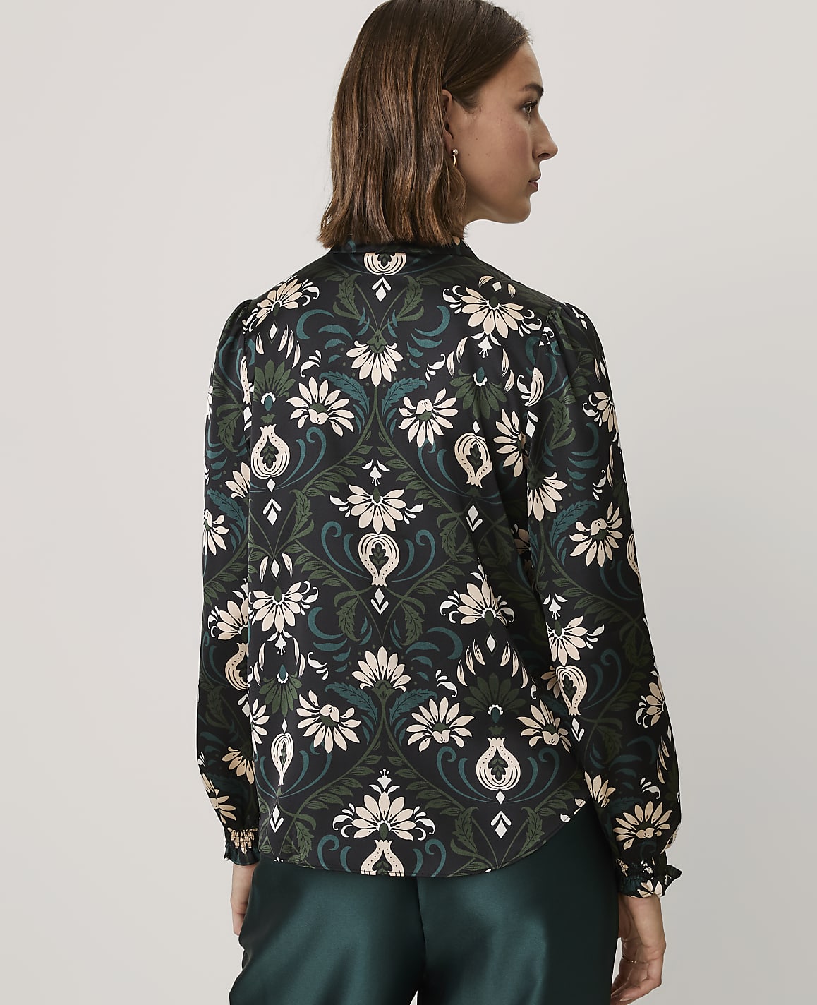 A woman wearing a floral print blouse and dark green satin trousers stands against a plain light gray background.