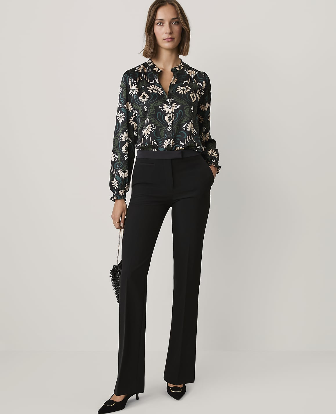 A woman wearing a patterned blouse and black trousers stands against a plain light grey background.