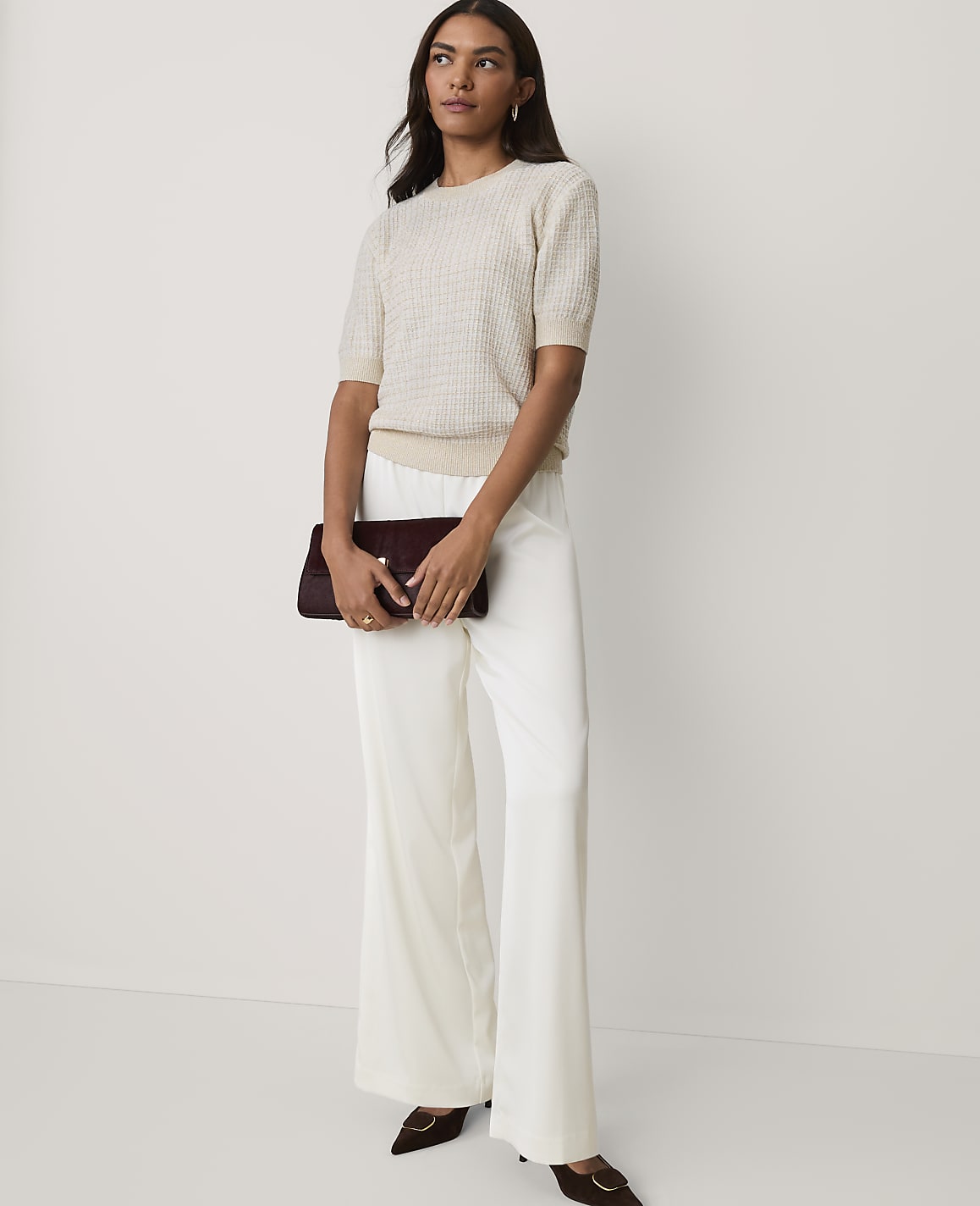 A woman wearing a cream-colored knit top and wide-leg white trousers holds a burgundy clutch against a plain white background.