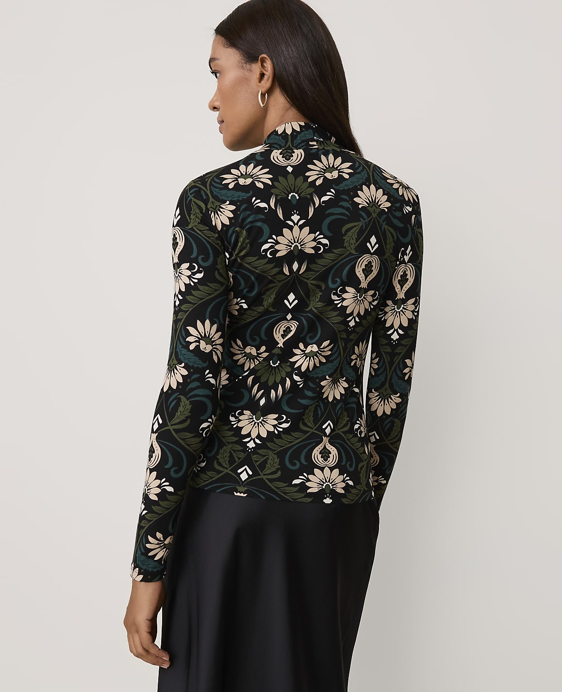 A woman wearing a floral patterned top and a black skirt stands against a plain light gray background.