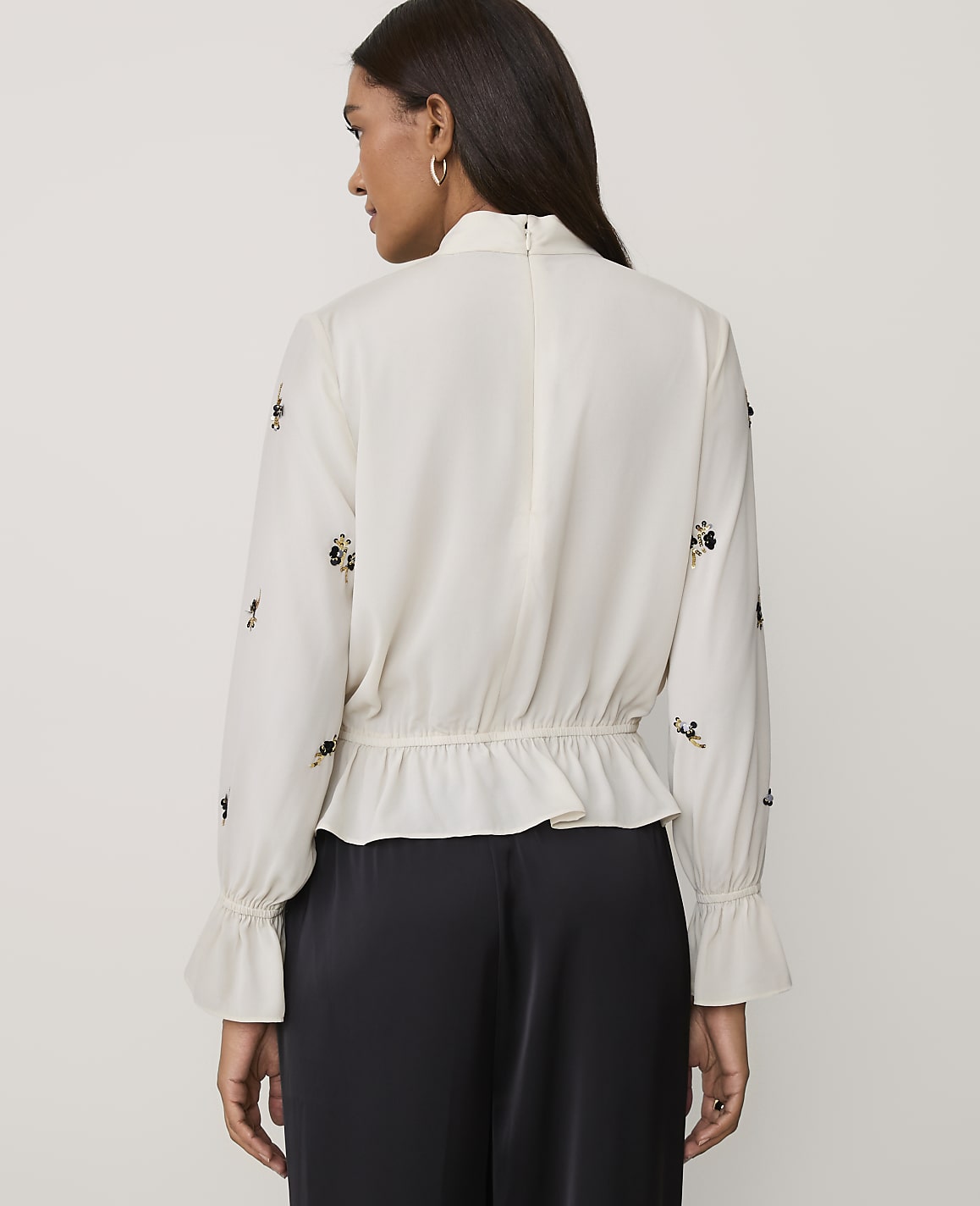 A woman stands with her back to the camera wearing a white embellished blouse and black trousers against a plain light grey background.