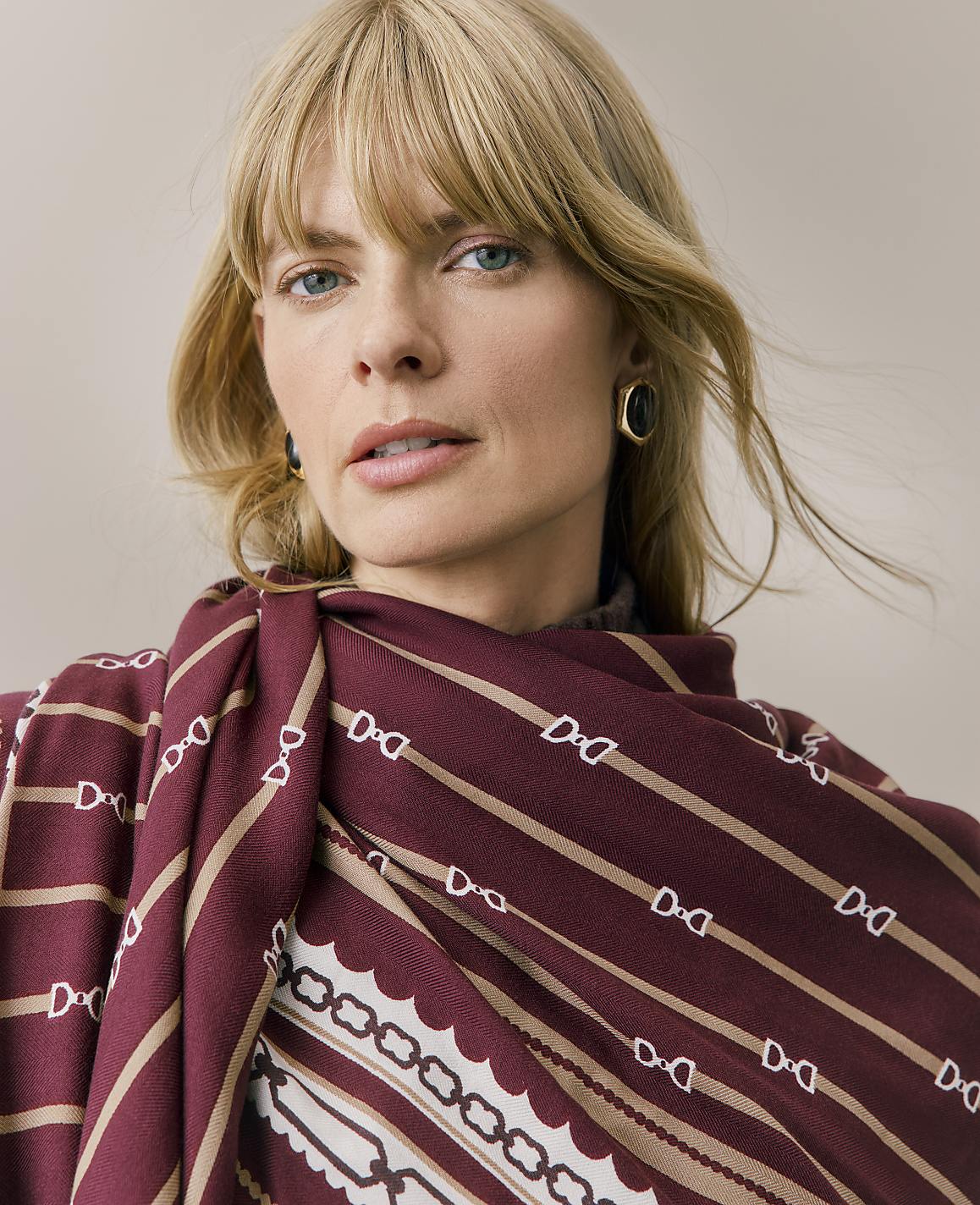 A blonde woman wearing a patterned burgundy and beige scarf and gold earrings poses against a plain, neutral background.