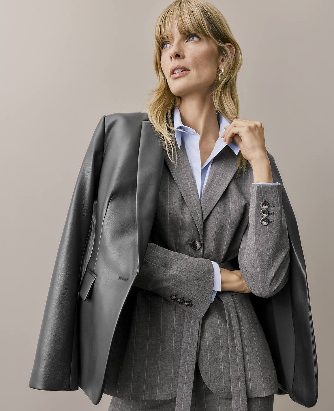 A blonde woman wearing a pinstriped grey suit and light blue shirt with a dark grey leather jacket draped over her shoulders stands against a plain beige background.