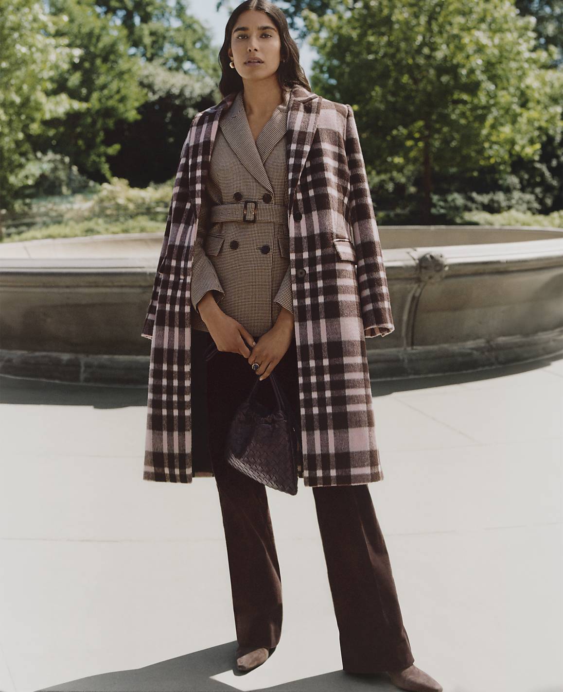 A woman wearing a plaid coat and brown trousers stands in the foreground before a stone fountain and lush green trees in the background.