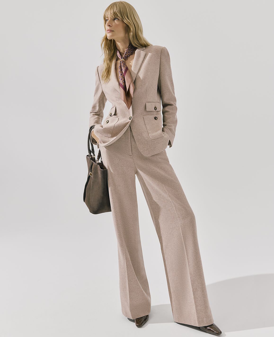 A blonde woman wearing a mauve power suit and a patterned scarf stands against a plain white background.